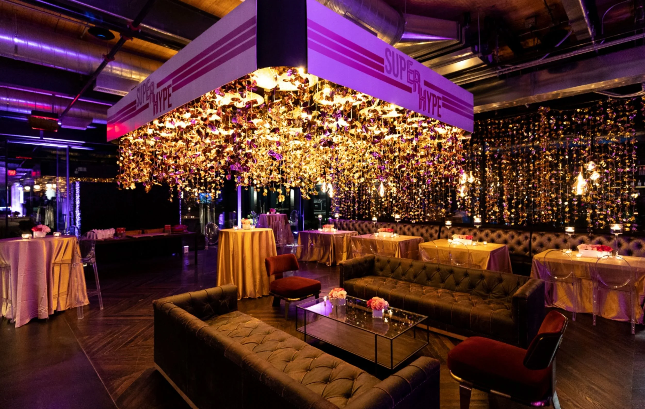 Luxurious event space with hanging floral decorations, tufted sofas, draped tables, and sign reading 'Super Hype'.