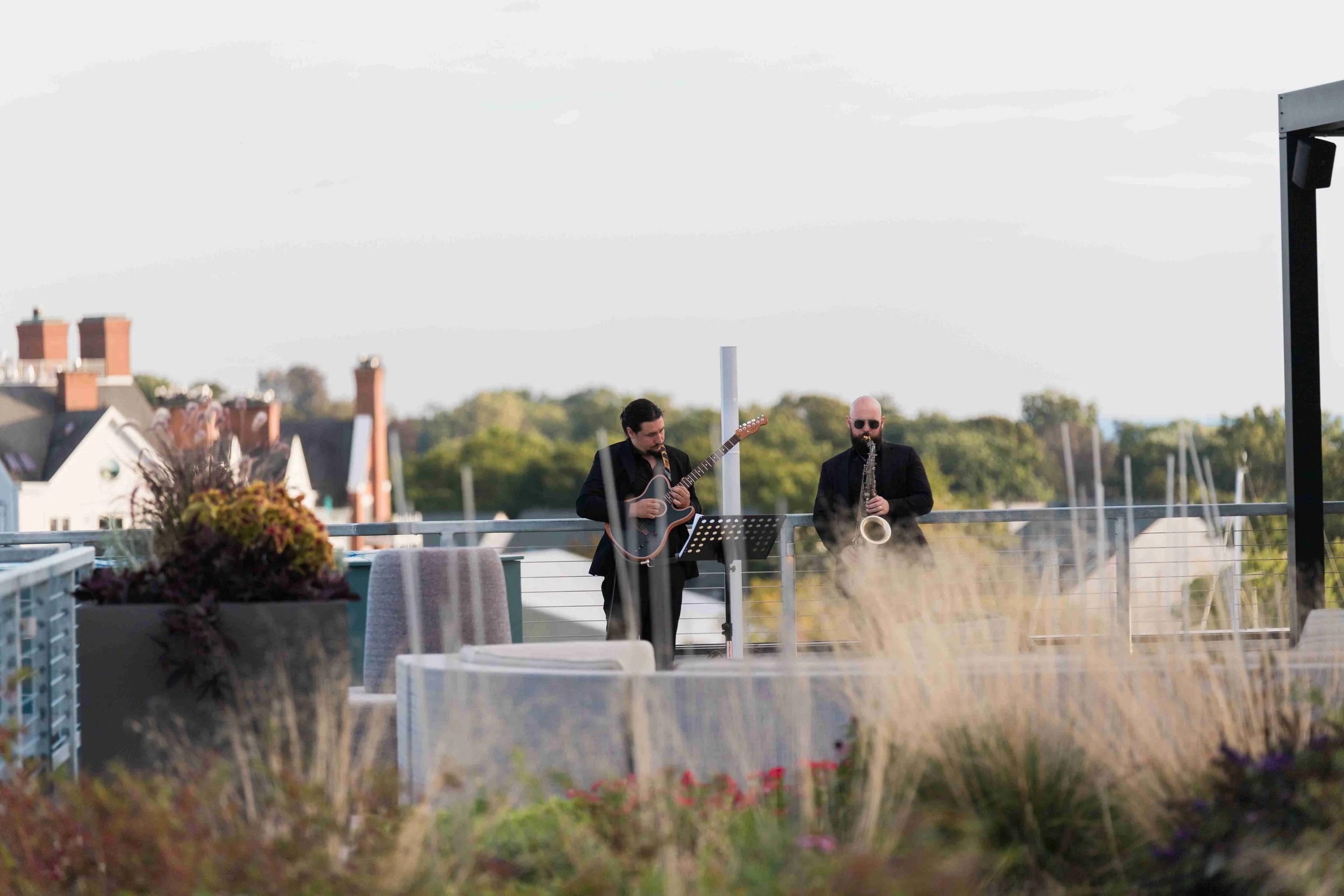 Two musicians, one playing an acoustic guitar and the other playing a saxophone, perform outdoors on a rooftop deck with a view of houses and trees in the background.