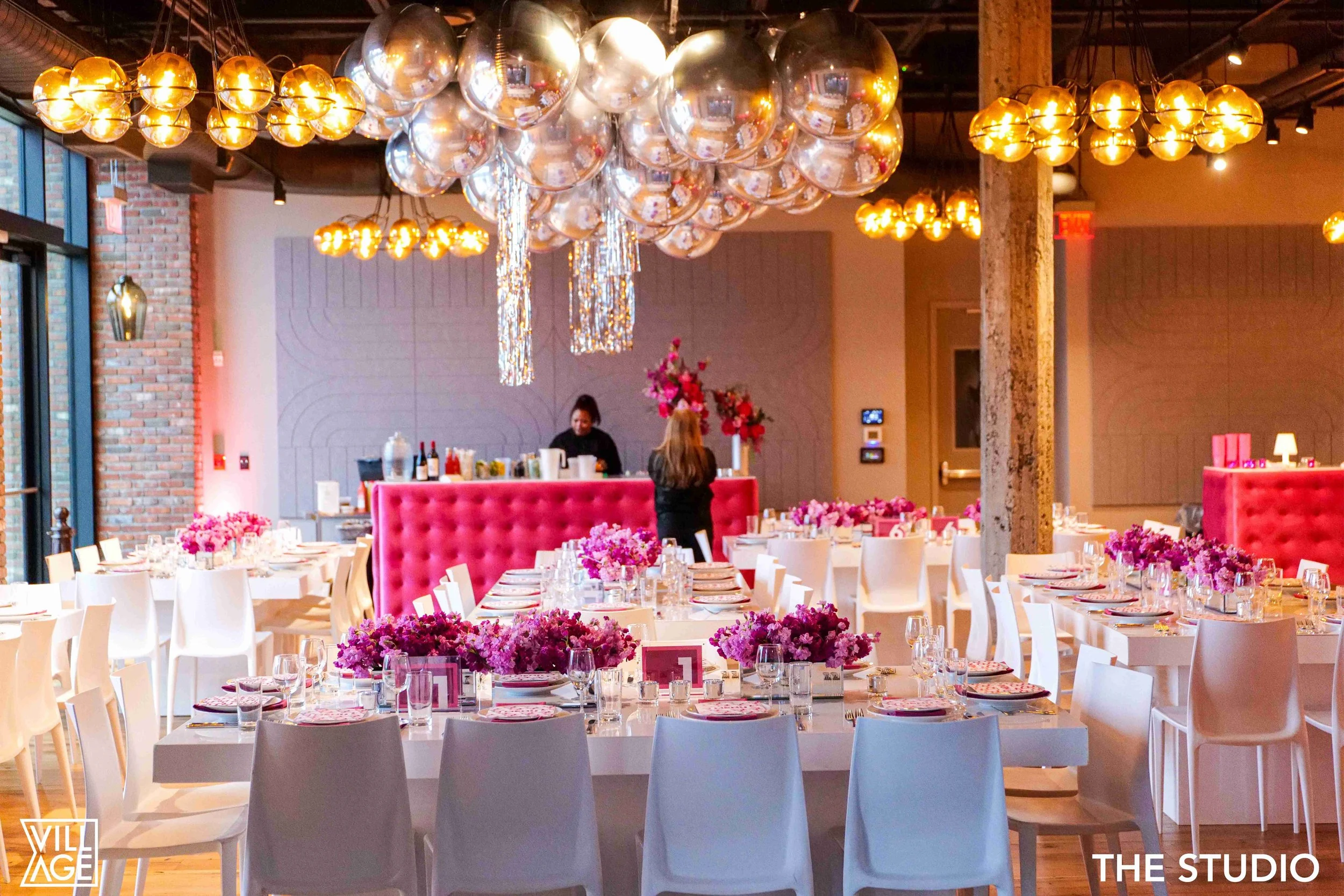 Interior of a decorated event space featuring pink floral centerpieces, white tables and chairs, a pink tufted partition, and large balloons and chandeliers hanging from the ceiling, with some staff preparing behind the bar.
