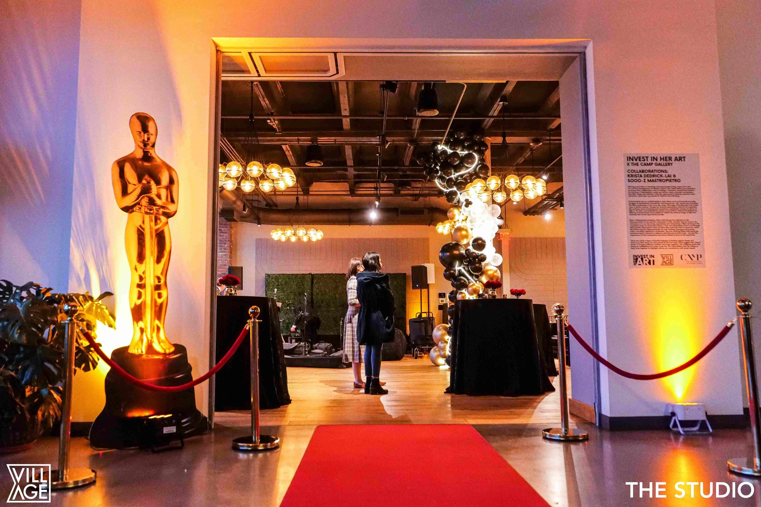 View of an art exhibit with a gold sculpture of an Oscar-like figure on the left and a balloon sculpture made of black, gold, and white balloons on the right. Two people are standing in the middle, viewing the art, with a small table covered in a bla