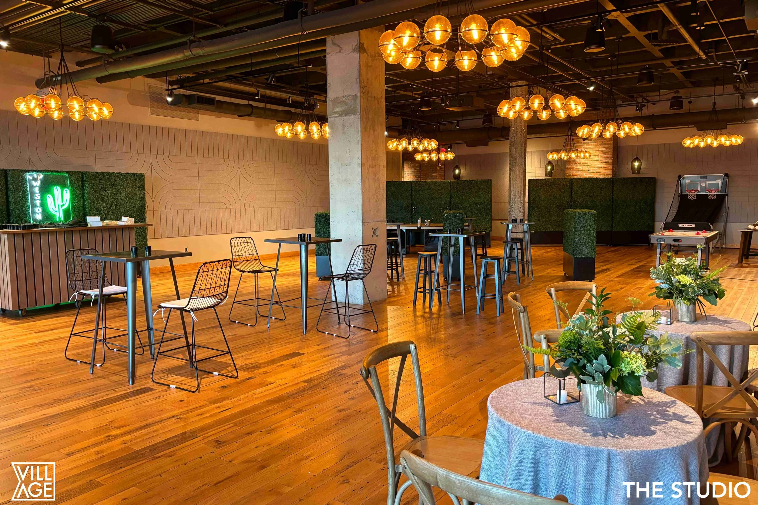 Interior of a modern event space with wooden floors, round tables with floral centerpieces, tall tables with bar stools, and a neon cactus sign. Warm lighting with multiple spherical chandeliers, a shuffleboard game, and privacy booths in the backgro