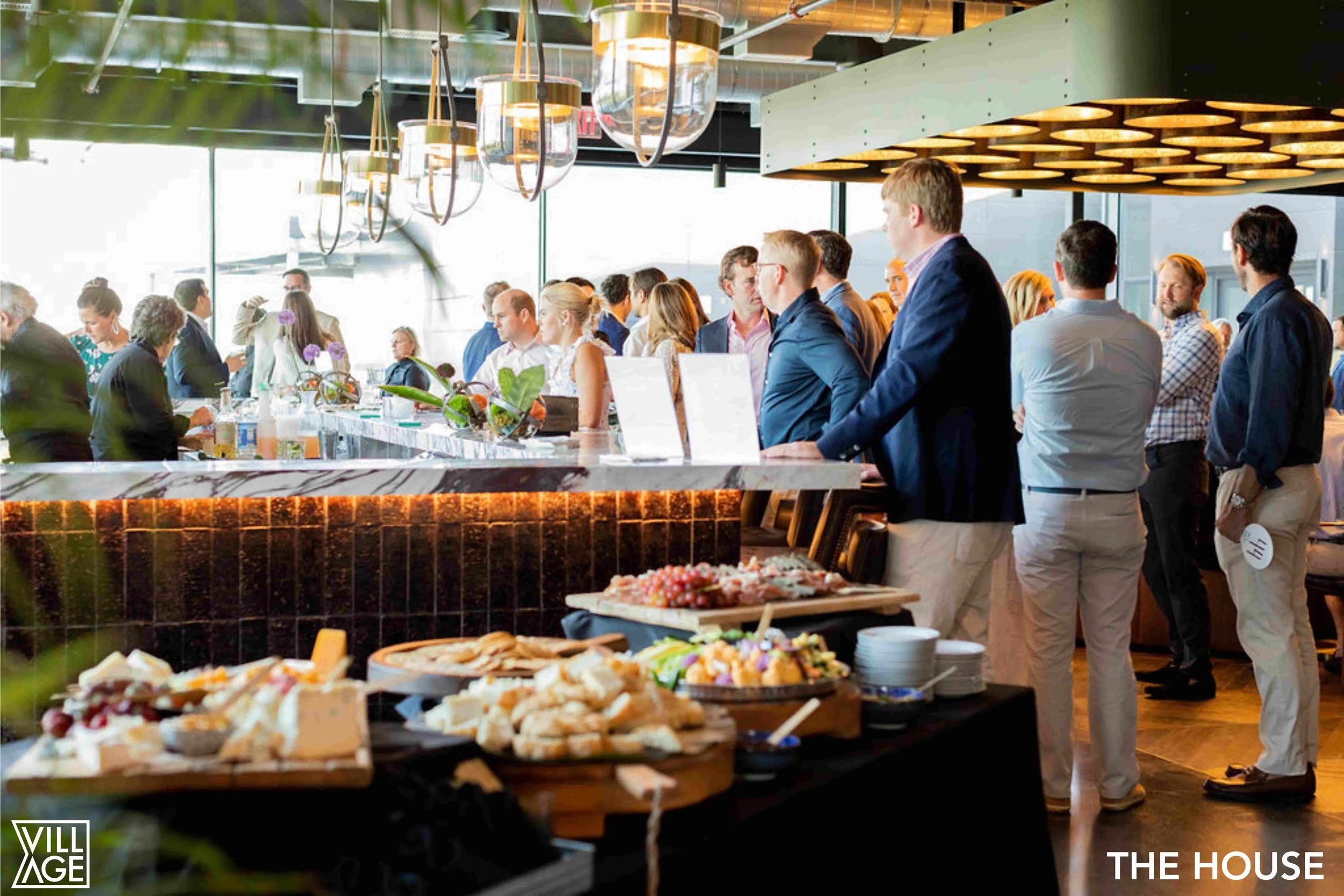People attending a networking event or conference in a modern, well-lit venue, gathered around a bar counter with refreshments and food.