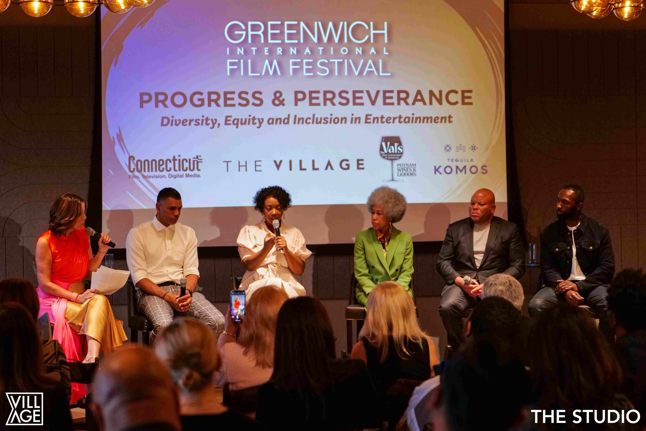 Six people seated on stage discussing at the Greenwich International Film Festival, with a large screen behind them displaying the festival's logo, theme 'Progress & Perseverance', and sponsors, including Connecticut, The Village, Val's, Tequila Komo