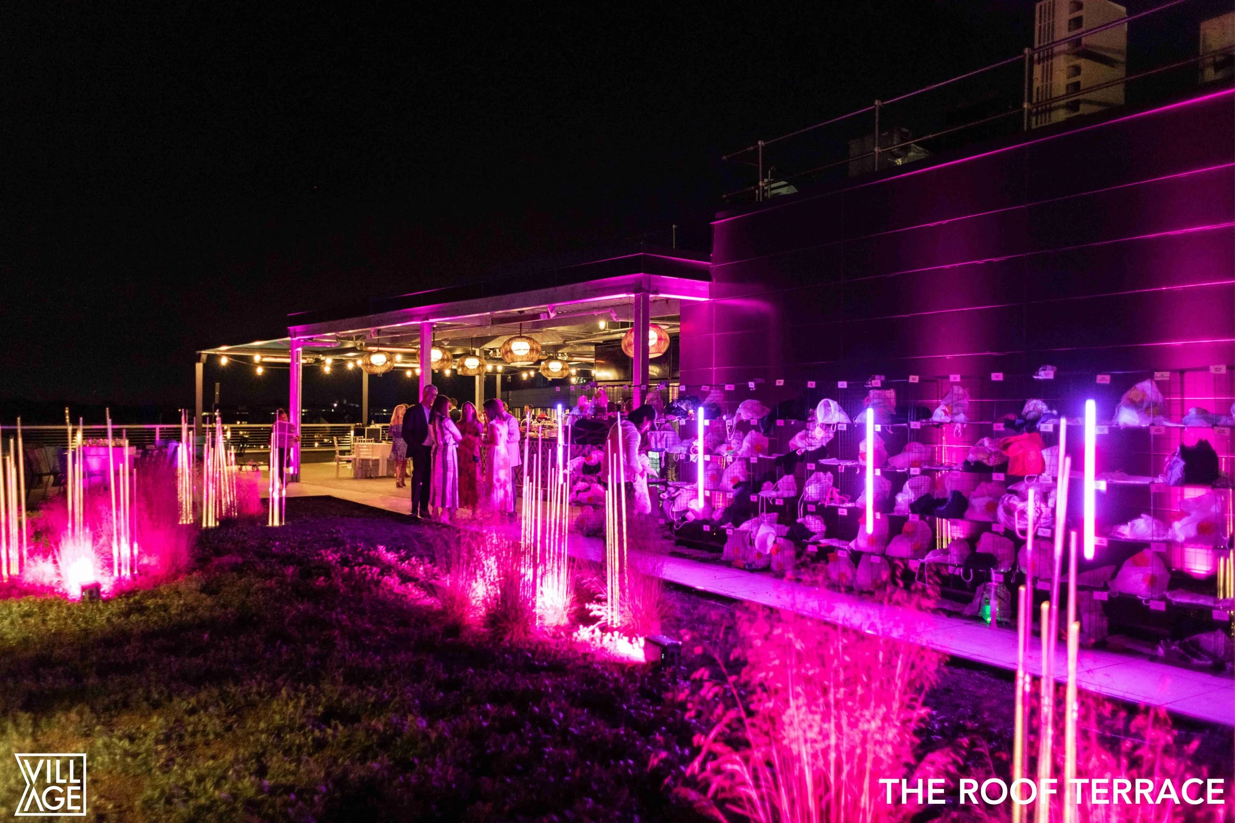 Nighttime rooftop terrace with purple and pink lighting, people socializing near a bar, illuminated decorative plants in the foreground, and city buildings in the background.