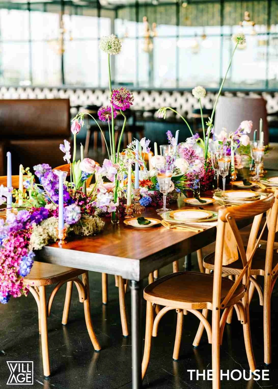 Elegant dining table decorated with colorful flowers in vases and tall candles, set with plates, glasses, and cutlery, in a bright, modern indoor space.