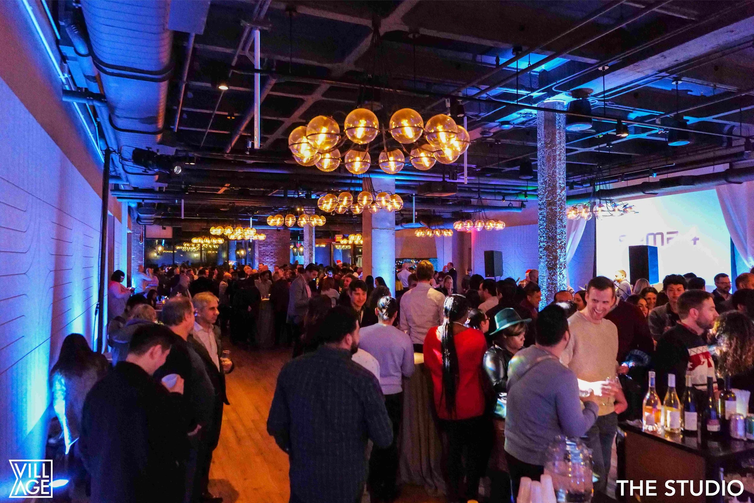 A crowded indoor event space with people socializing and drinking. The room is decorated with modern lighting, including hanging amber globes, and has a sleek, contemporary design with exposed ceiling pipes and brick columns. The atmosphere is lively