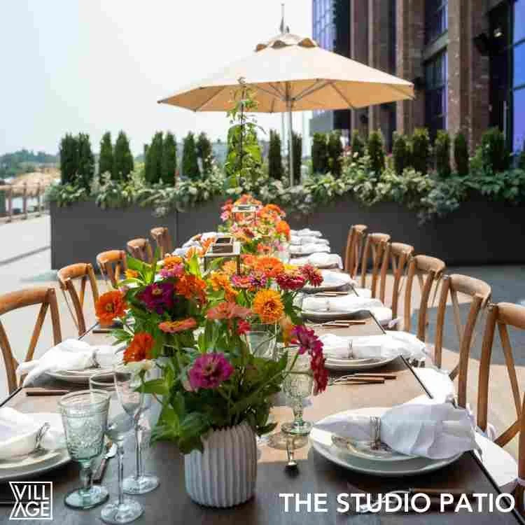Outdoor dining table on a patio decorated with vibrant flower arrangements under a large beige umbrella, with place settings and glasses arranged for a meal.