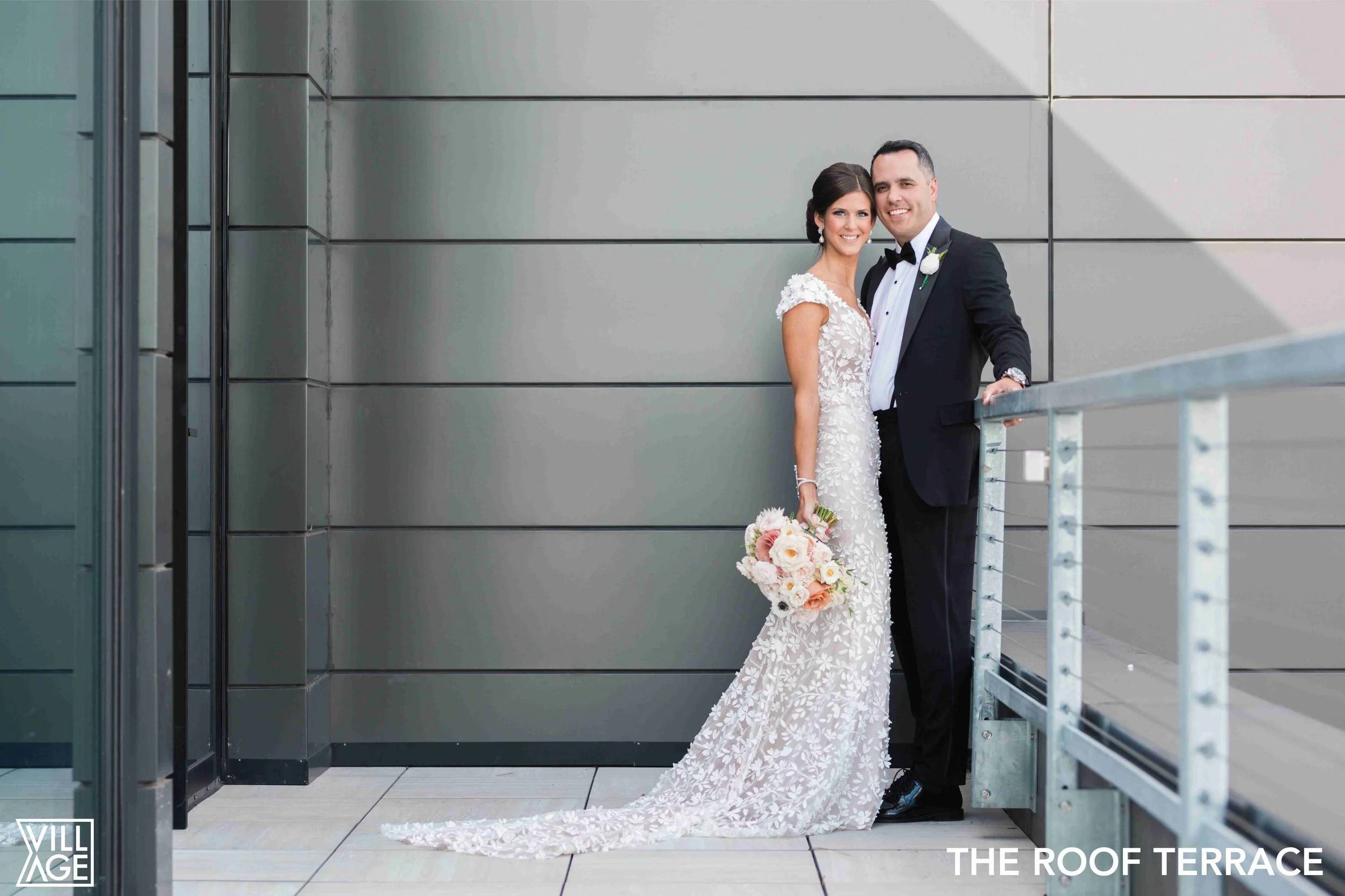 A newlywed couple in wedding attire standing on a modern roof terrace, with the bride holding a bouquet of flowers and the groom smiling next to her, against a sleek metal wall background.