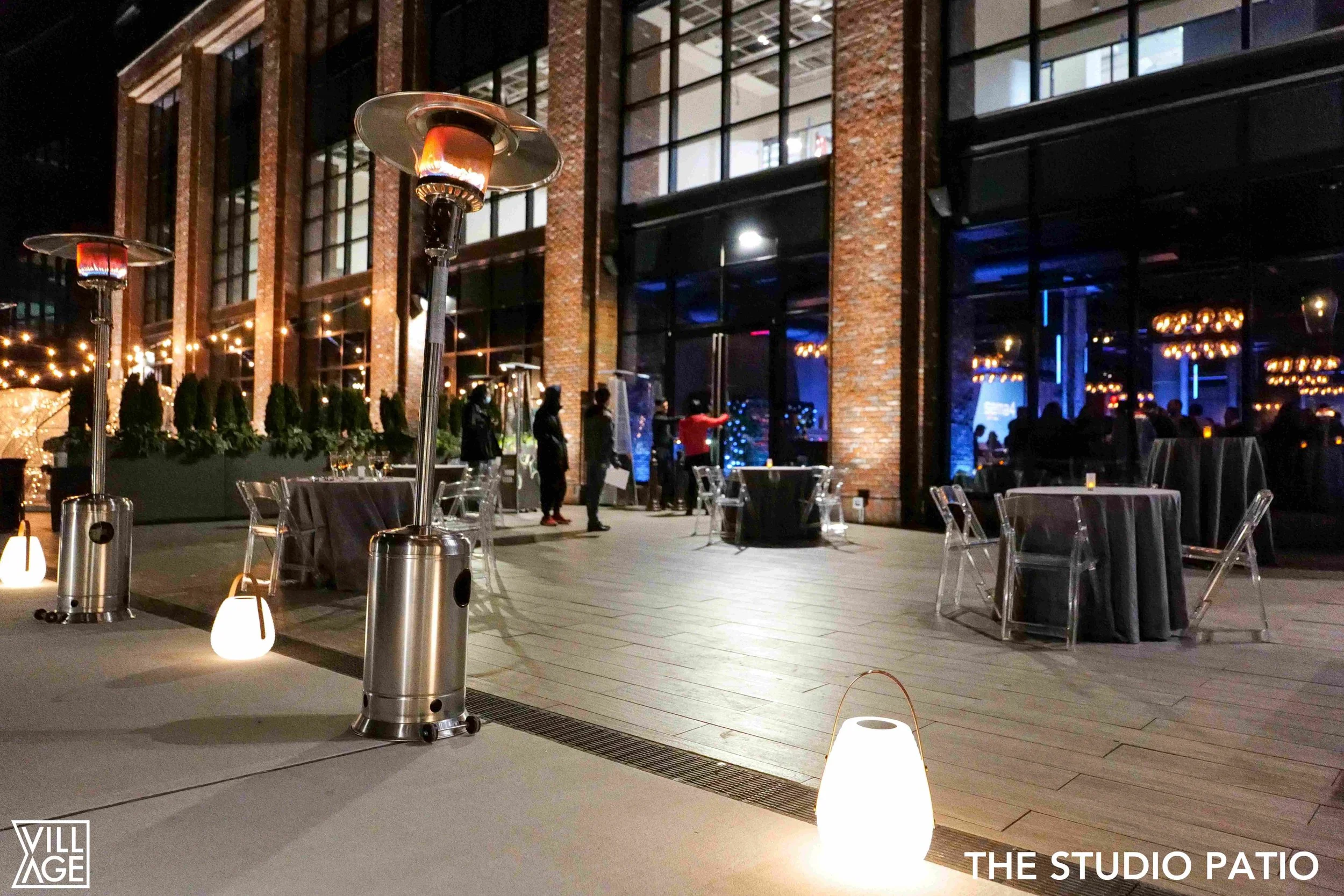 Outdoor patio at night with tables, chairs, and tall patio heaters. String lights and interior lights visible through large windows in a brick building. People are seen standing and sitting inside and outside.