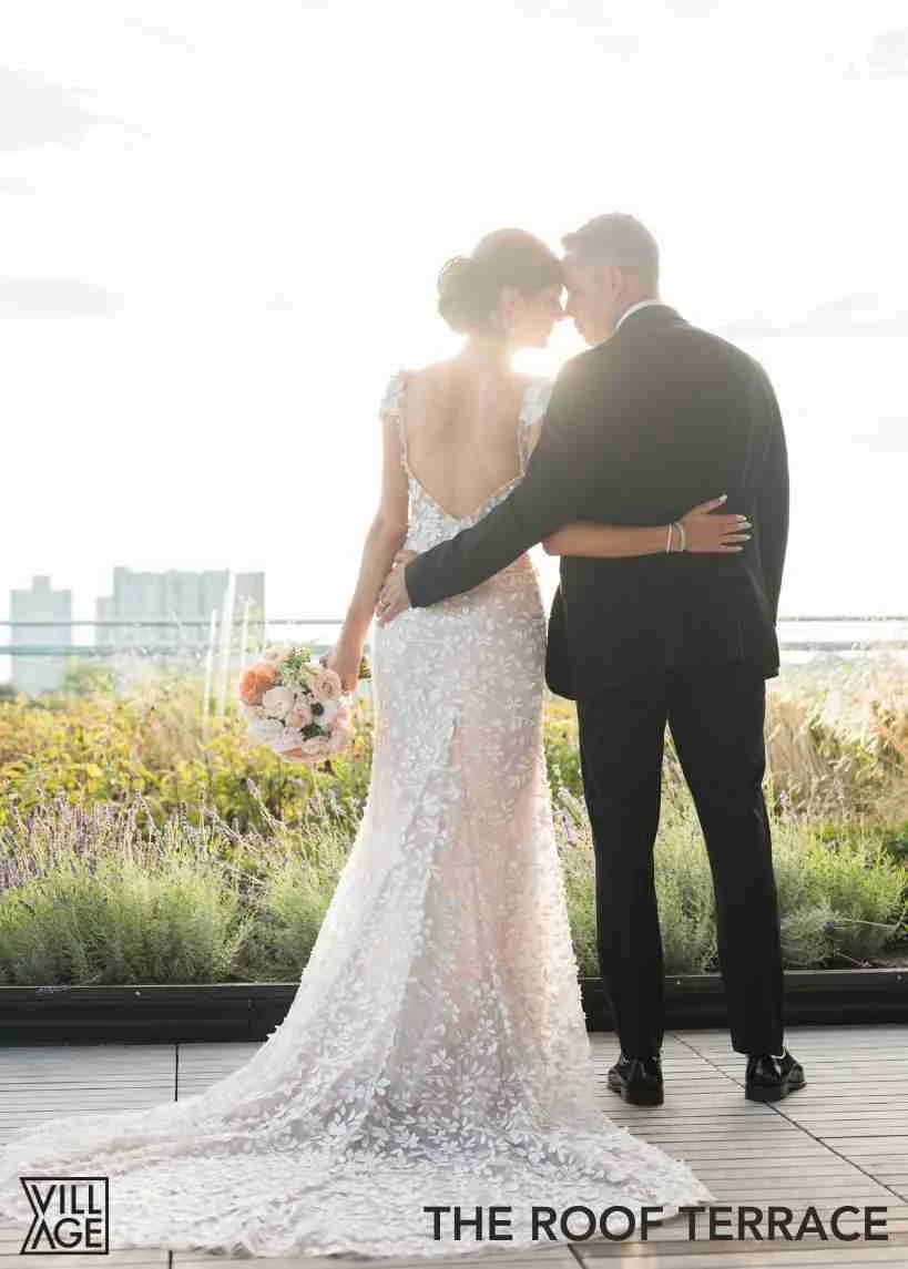 A newlywed couple, dressed in wedding attire, stands close together on a rooftop terrace. The bride holds a bouquet of flowers, and they are backlit by bright sunlight, creating a silhouette effect. The background features a cityscape with tall build