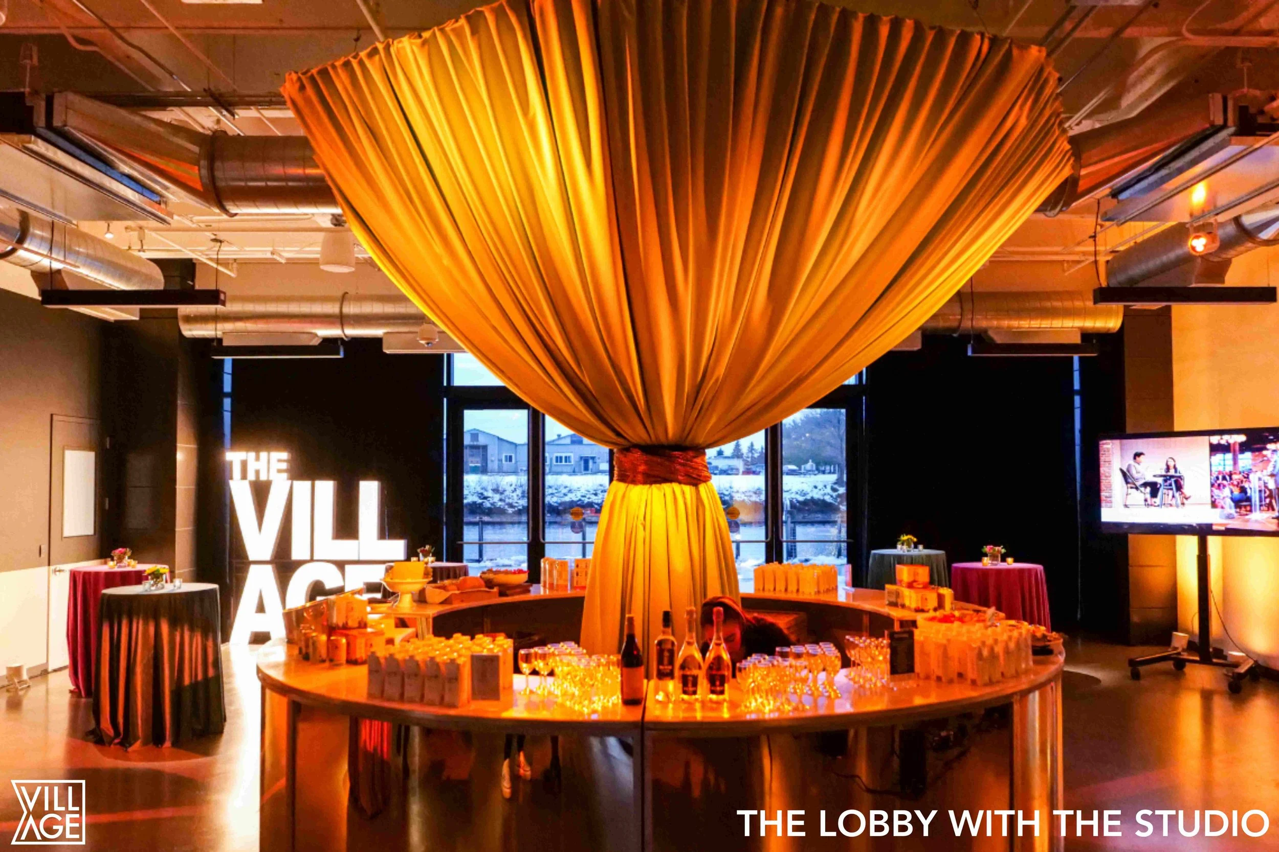 Interior of a modern event space with a round table displaying bottles and glasses, a large yellow fabric installation hanging from the ceiling, and a sign reading 'THE VILLAGE' in illuminated letters on the wall in the background.