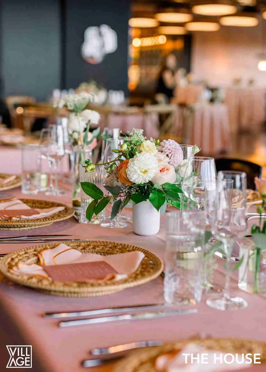 A table setup with a pink tablecloth, wicker placemats, pink napkins, silverware, clear glassware, and a centerpiece of white and pink flowers in a white vase, in a restaurant or event space with warm lighting.