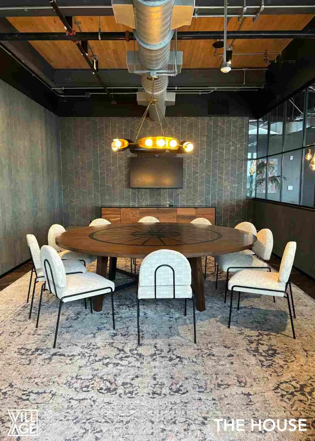 Modern conference room with a round wooden table, ten white cushioned chairs, a textured dark wall with an overhead chandelier light fixture, a wall-mounted television, and glass windows on the right side.