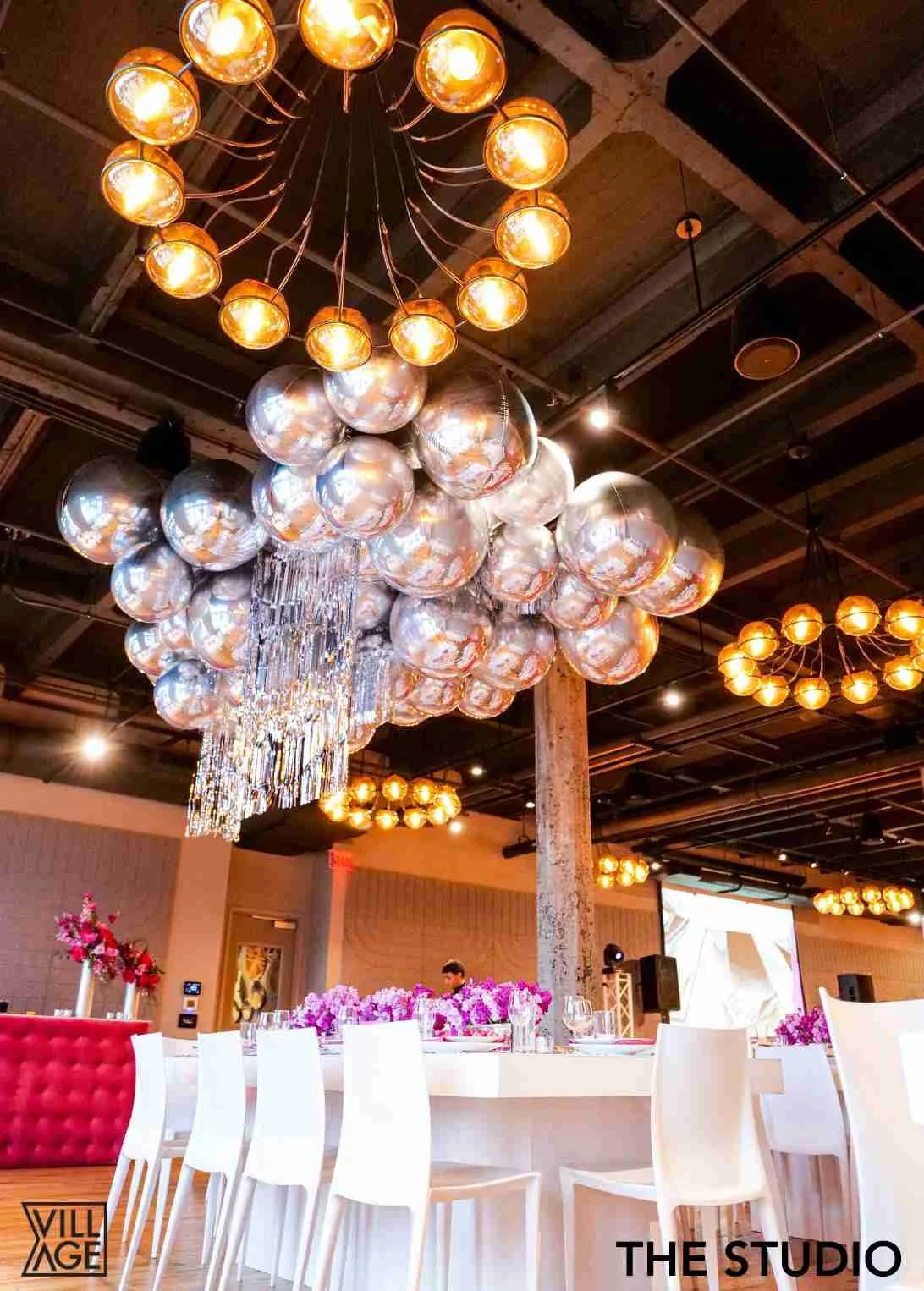 Elegant event space decorated with large, modern chandeliers made of multiple spherical metallic and glass elements, with a long white table adorned with pink floral centerpieces, surrounded by white chairs, and a projection screen in the background.