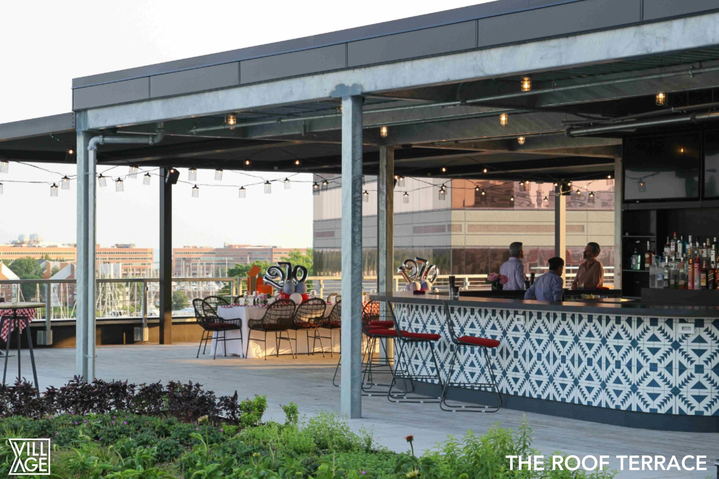Rooftop terrace bar with string lights, bar area with three people, decorated tables, and a city view in the background.