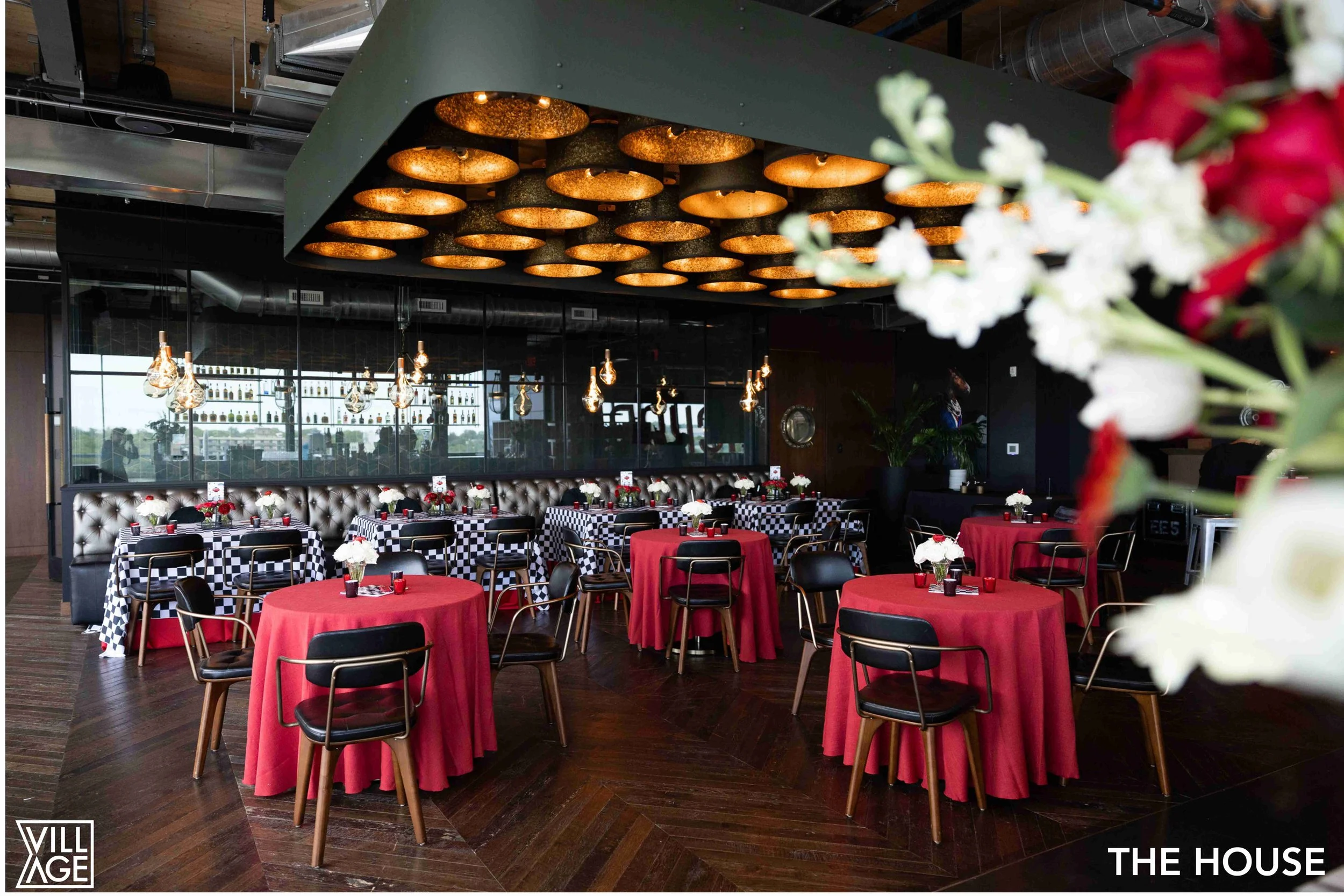 Restaurant interior with round tables covered in red tablecloths, black chairs, floral centerpieces, black and white checkered and silver tufted booth seating, and modern hanging light fixtures.
