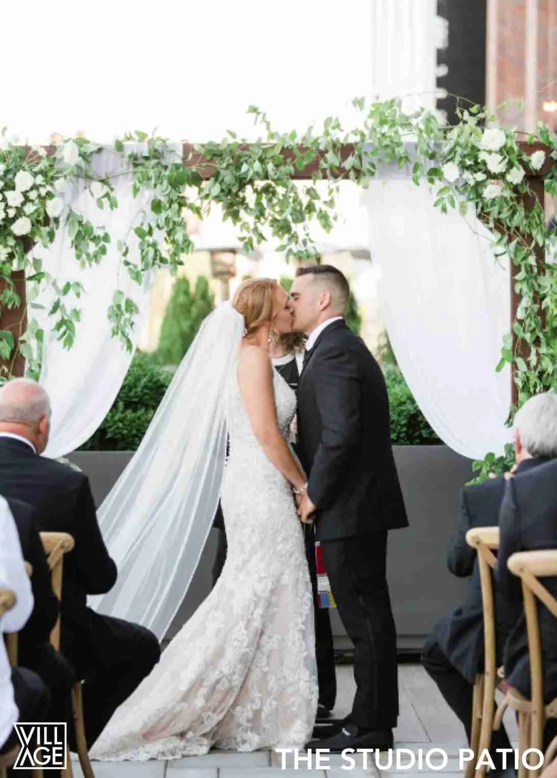 A bride and groom kiss during their outdoor wedding ceremony under a floral arch, with seated guests in the foreground.