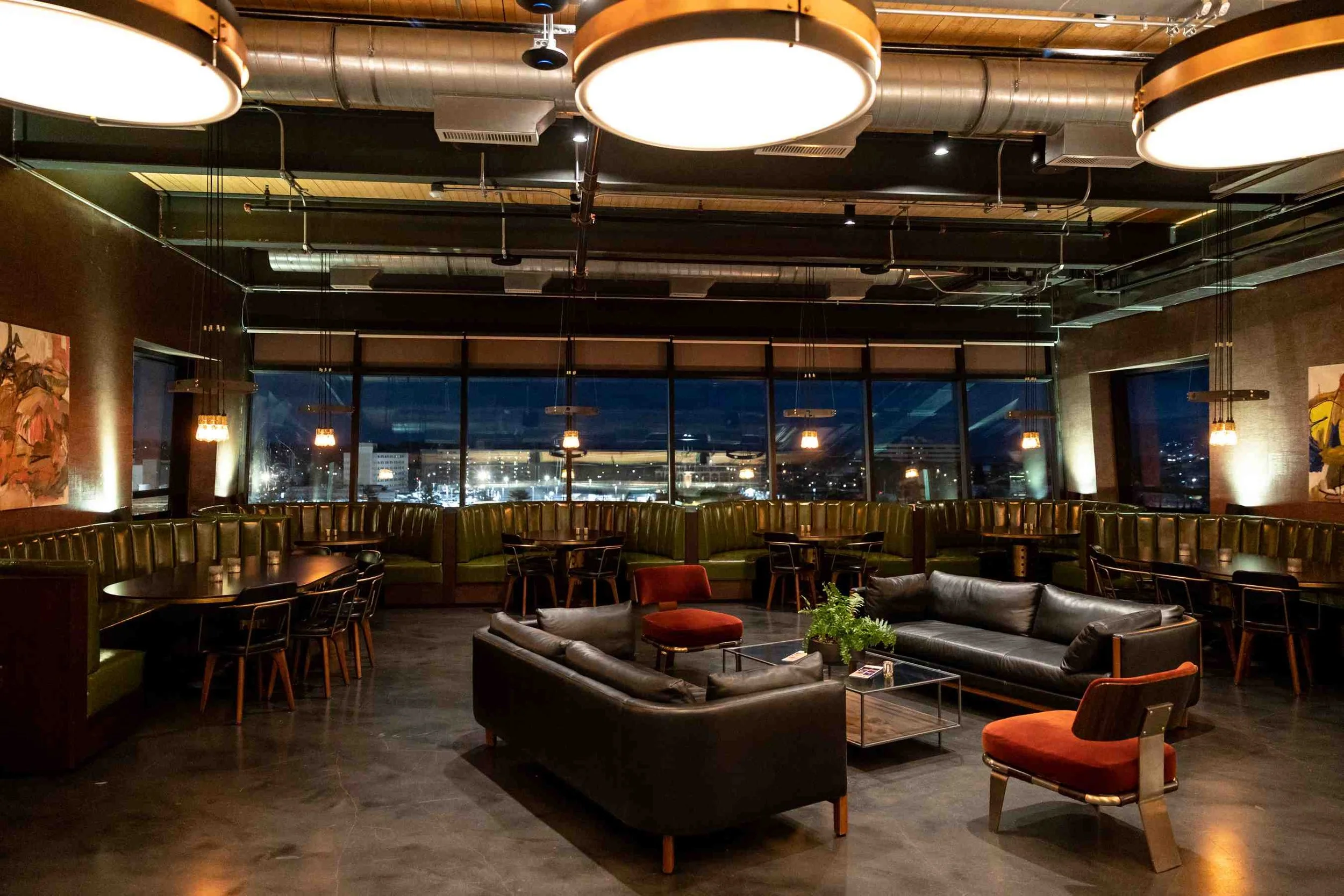 Modern lounge area with green leather seating, wooden chairs, a potted plant, pendant light, and a bar with hanging lights and a mural in the background.