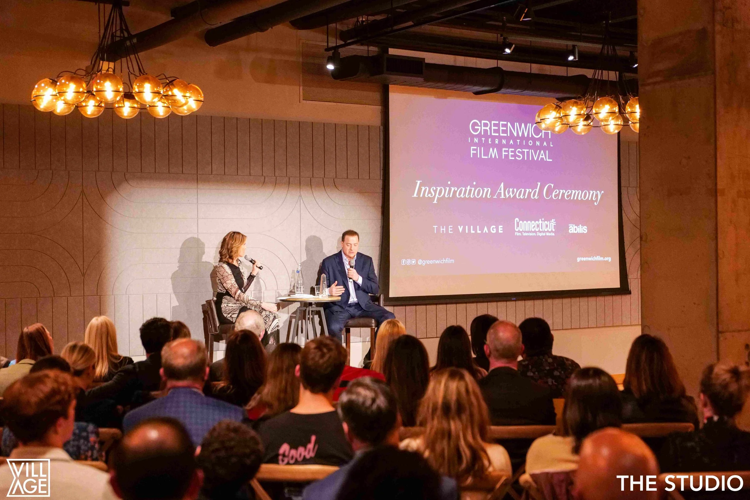 An award ceremony at Greenwich International Film Festival with a woman and a man sitting on stage, speaking into microphones in front of an audience. A large screen behind them displays the event's title and sponsors.