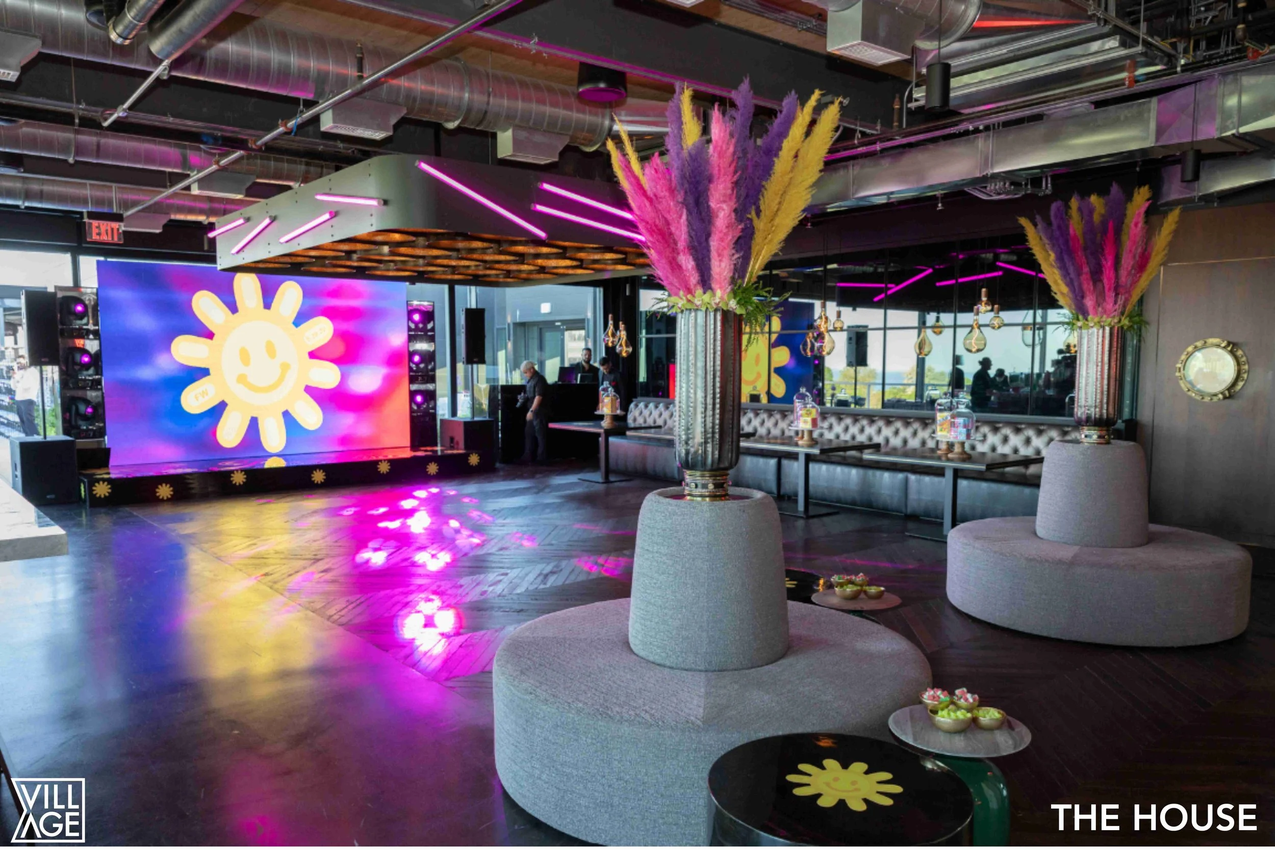 Interior of a modern event space with a large LED screen displaying a yellow smiley face flower, surrounded by colorful lighting and decor, including tall vases with pink, purple, and yellow feathered flowers, gray cushioned seating, and small bowls 