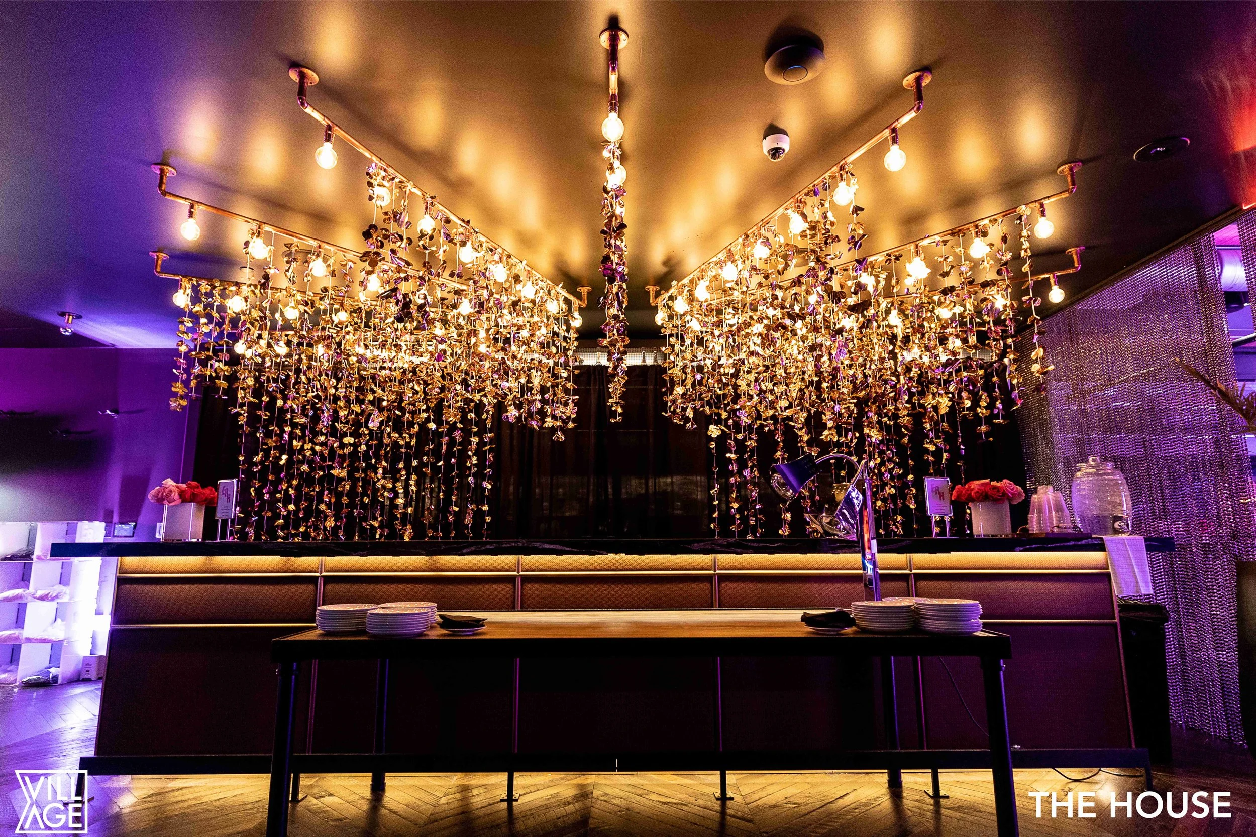 Interior of a stylish event space with a decorated ceiling featuring hanging string lights and floral decorations, a bar with plates and a lamp, and purple lighting accents.