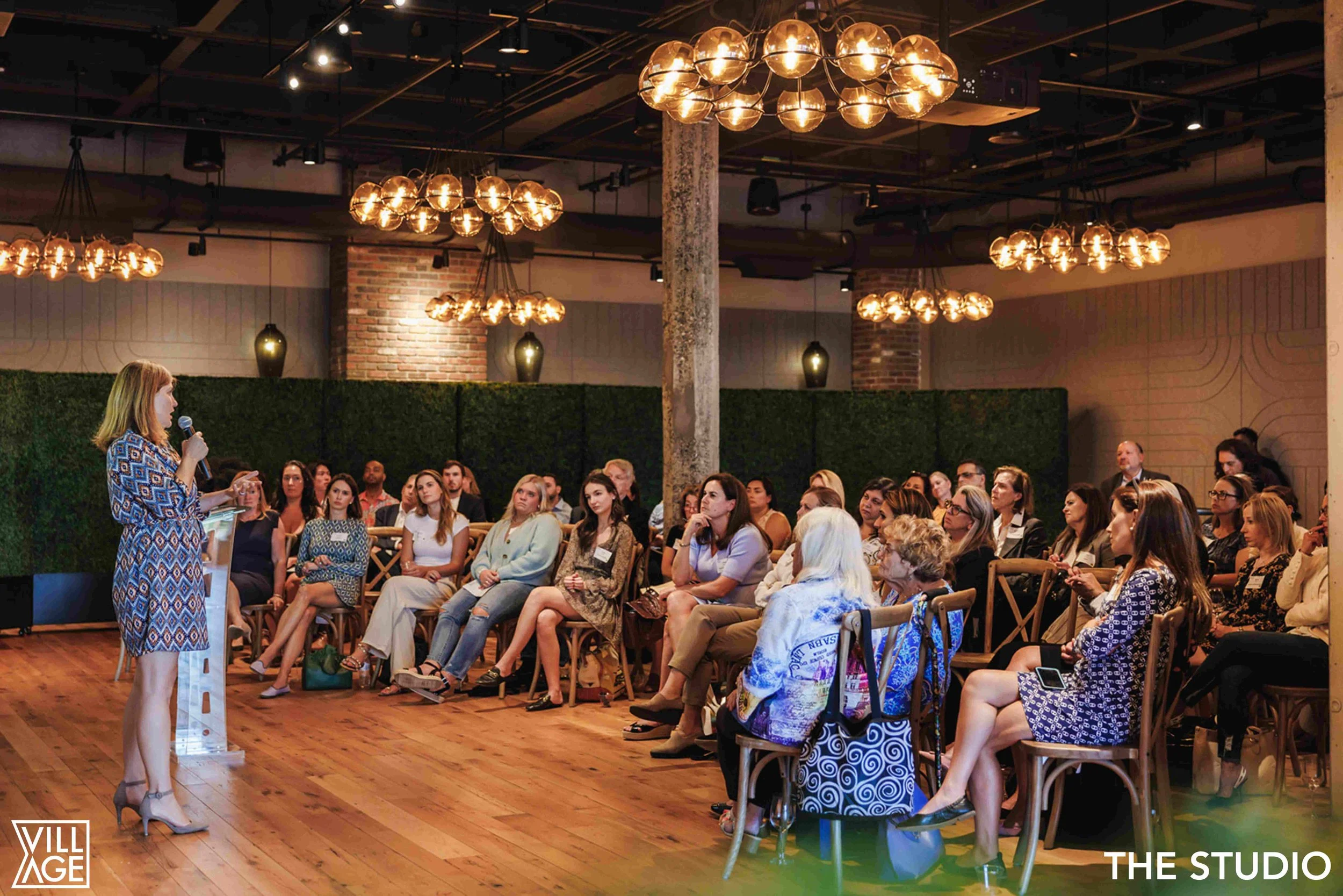 A woman in a patterned dress presenting to an audience in a conference or seminar room with wooden floors, modern lighting, and exposed brick walls.