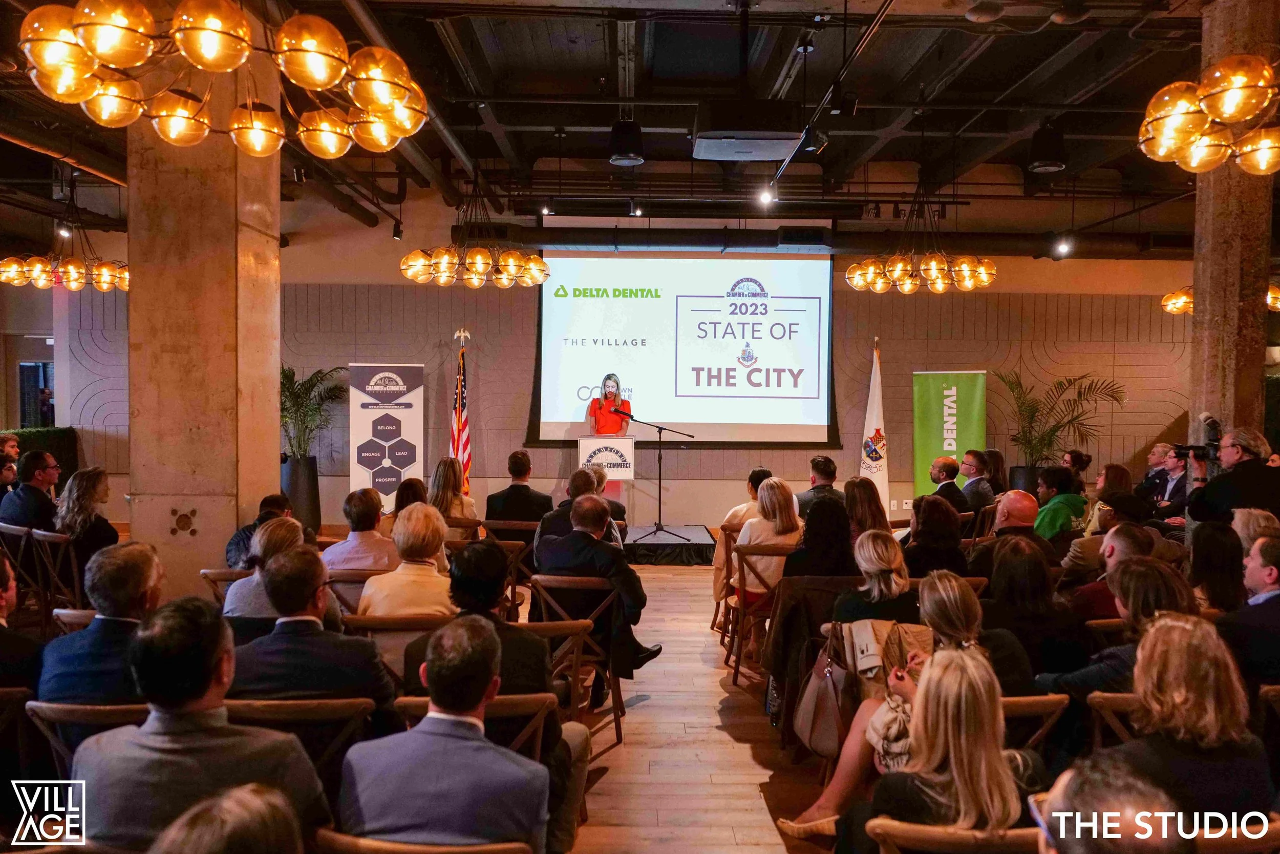A woman in a red dress is speaking at a podium during a conference titled 'State of the City 2023'. The event is held in a room with warm lighting and an audience seated in chairs facing the stage. There are American flags on either side of the stage