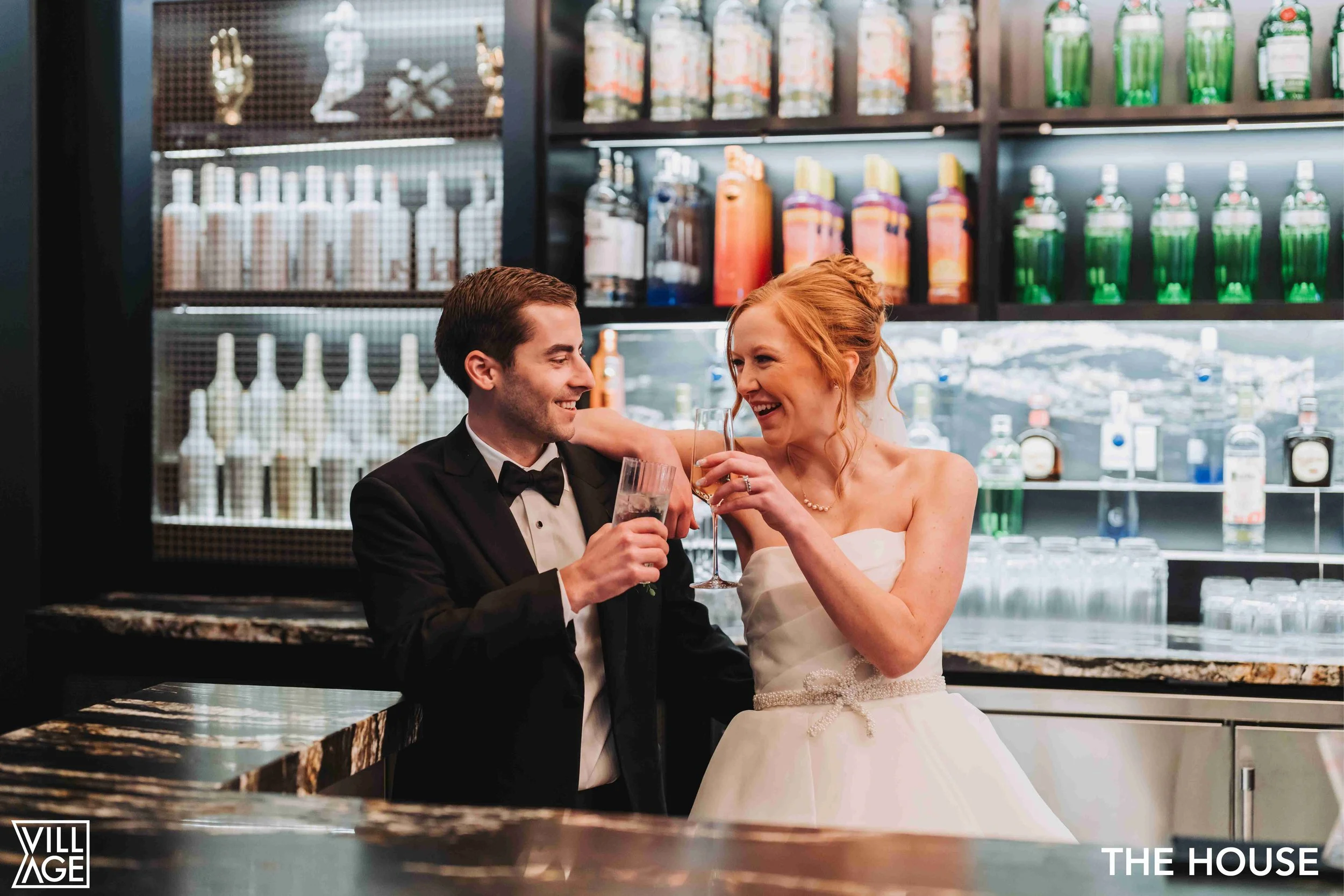 A man and woman, dressed in formal attire, celebrating with drinks in a modern bar with shelves stocked with bottles and glasses.