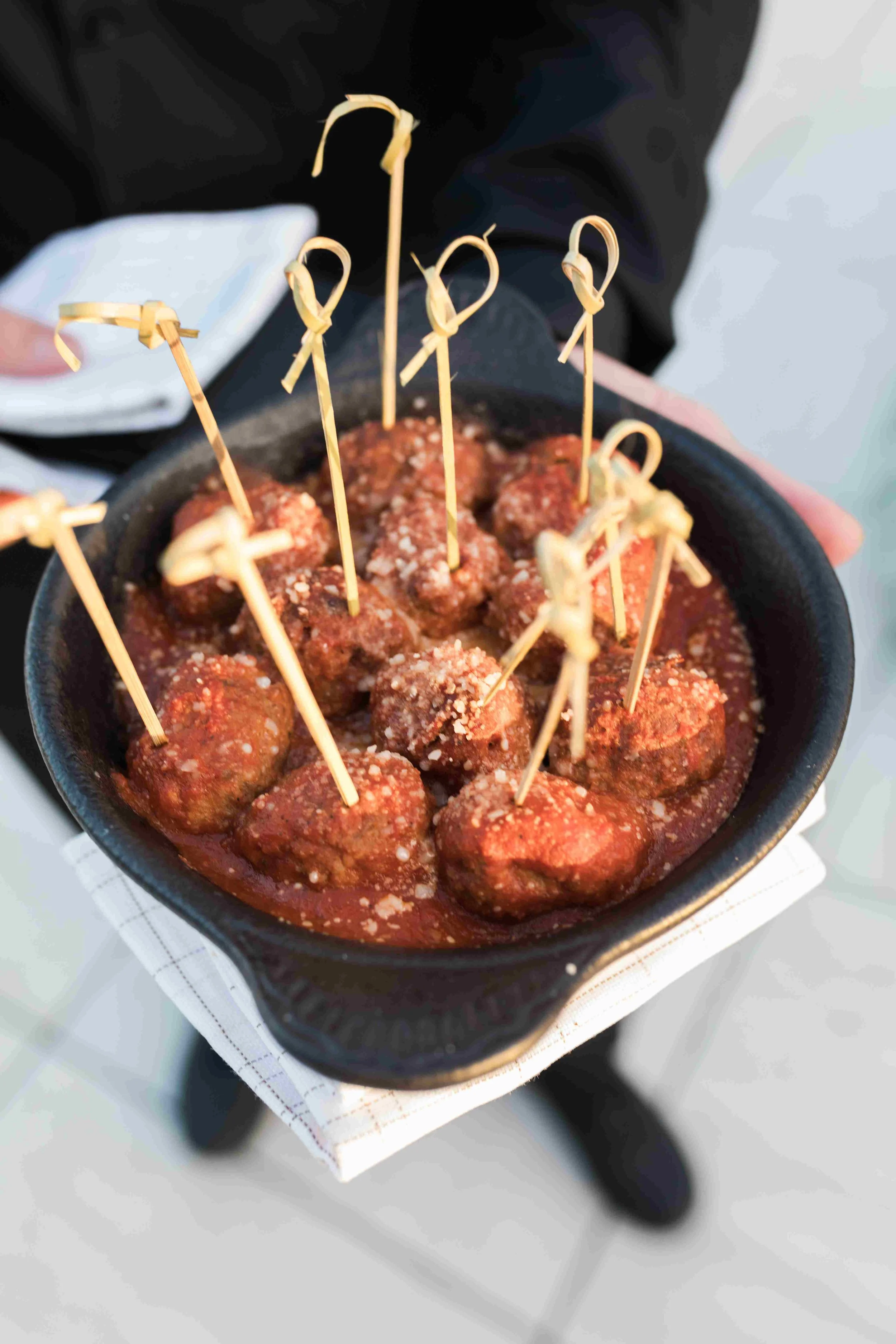 A bowl of cocktail meatballs with toothpicks, served in a red sauce and topped with grated cheese, held by a person wearing a black jacket.