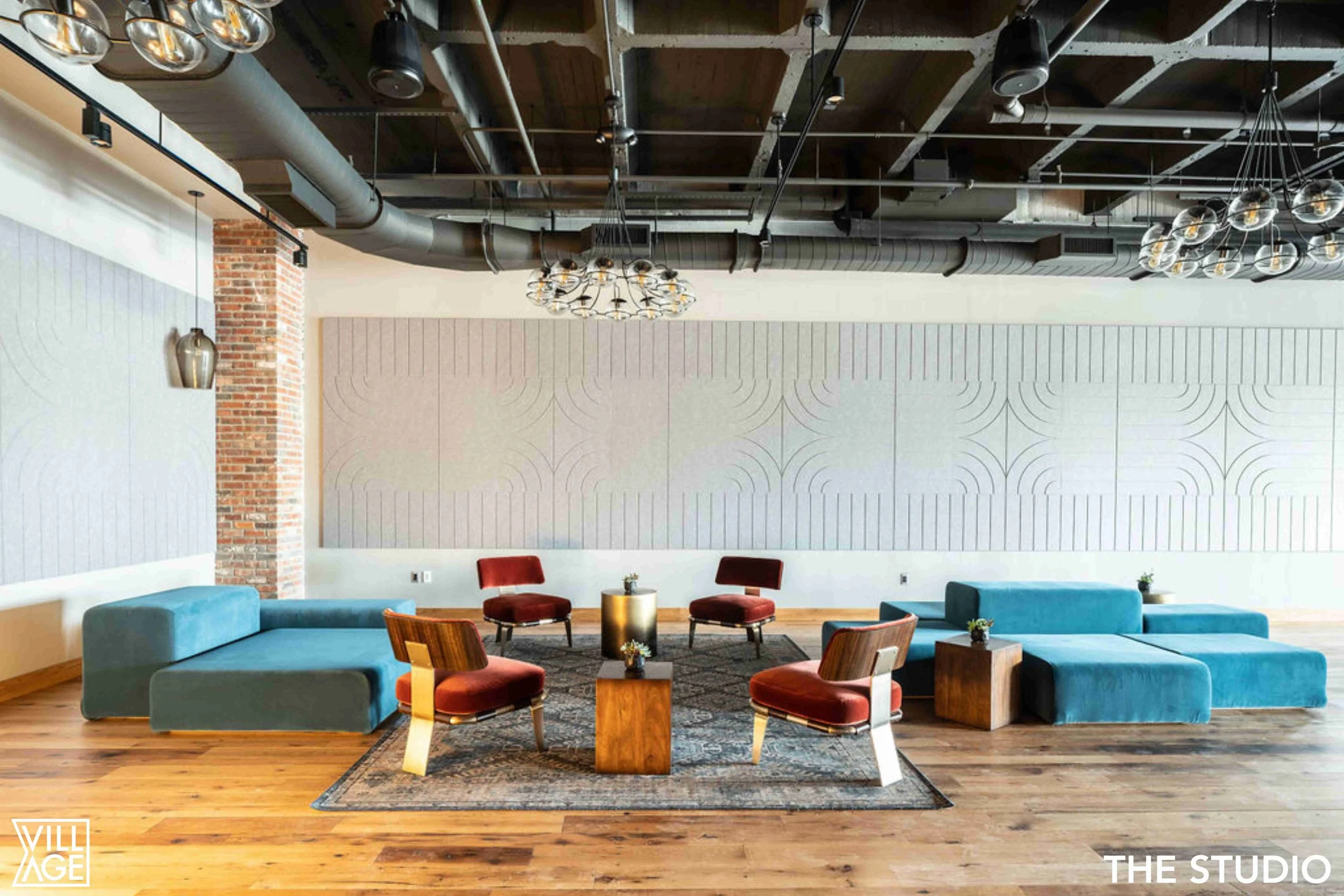 Modern lounge area with blue sectional sofa, four red velvet chairs, wooden side tables, potted plants, brick and white wall decor, hardwood floor, and industrial ceiling with exposed ductwork and black track lighting.