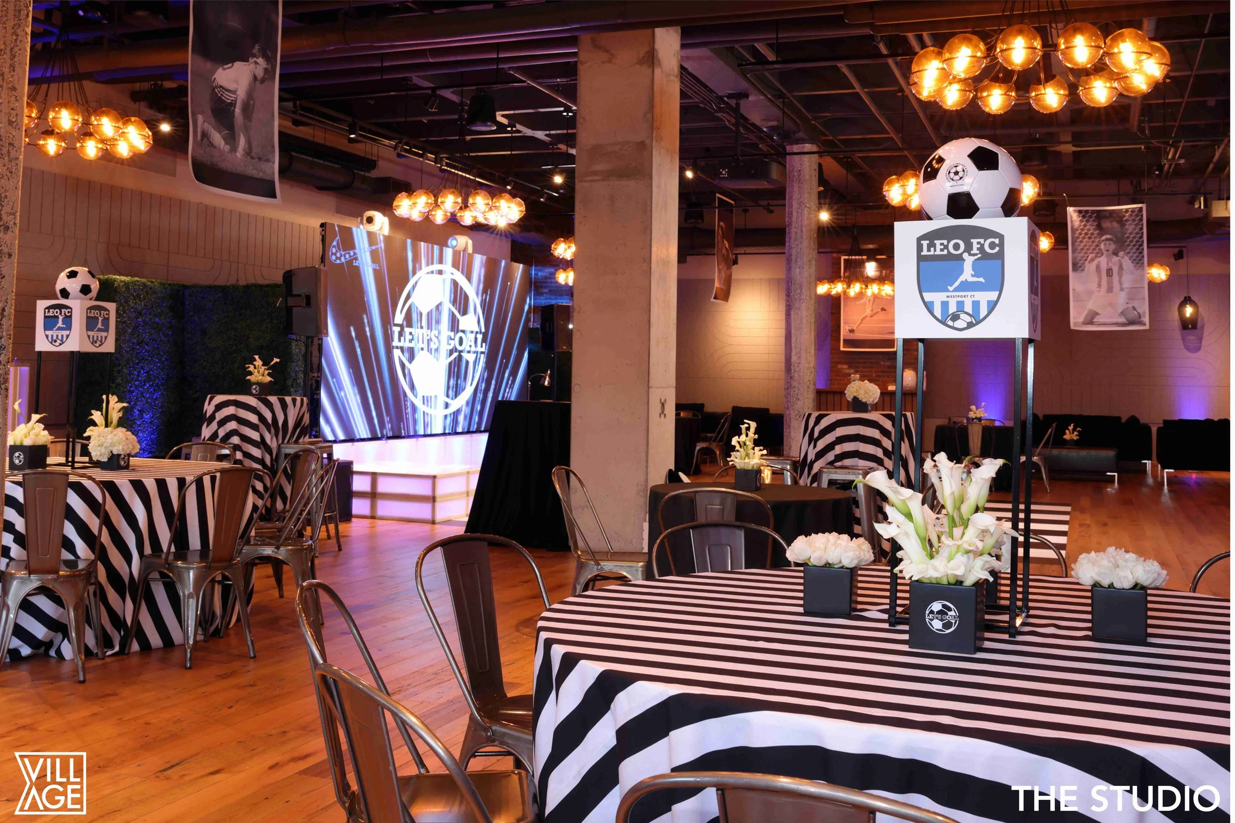Decorated event space with tables covered in black and white striped tablecloths, floral centerpieces, large soccer-themed balloons, and lighting fixtures, featuring a big screen displaying 'LEO FC' logo.