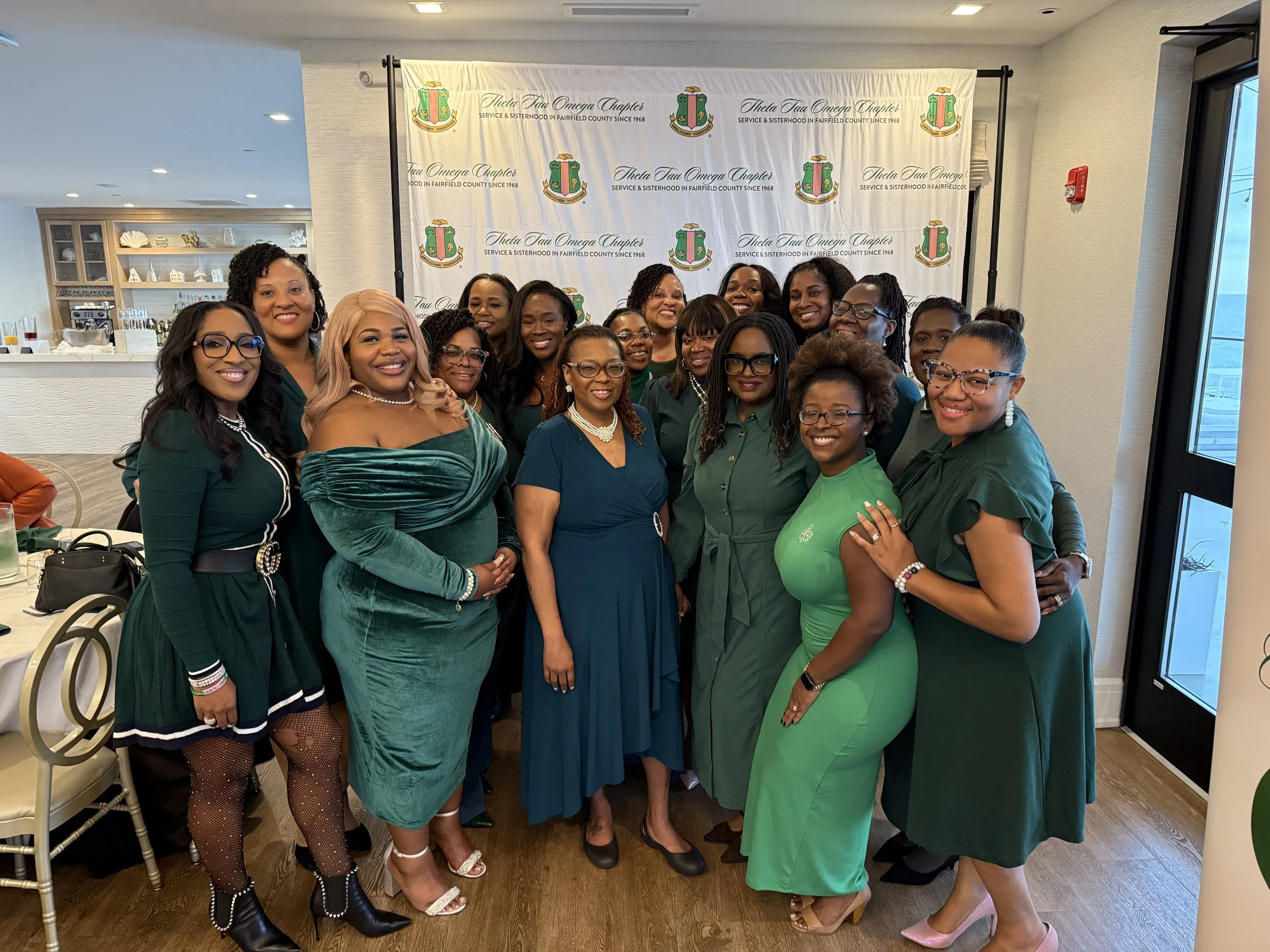 Group of women wearing green dresses and outfits attending a social event in a banquet hall with a backdrop that reads 'Theta Tau Omega Chapter Service & Sisterhood in Fairfield County Since 1968'.