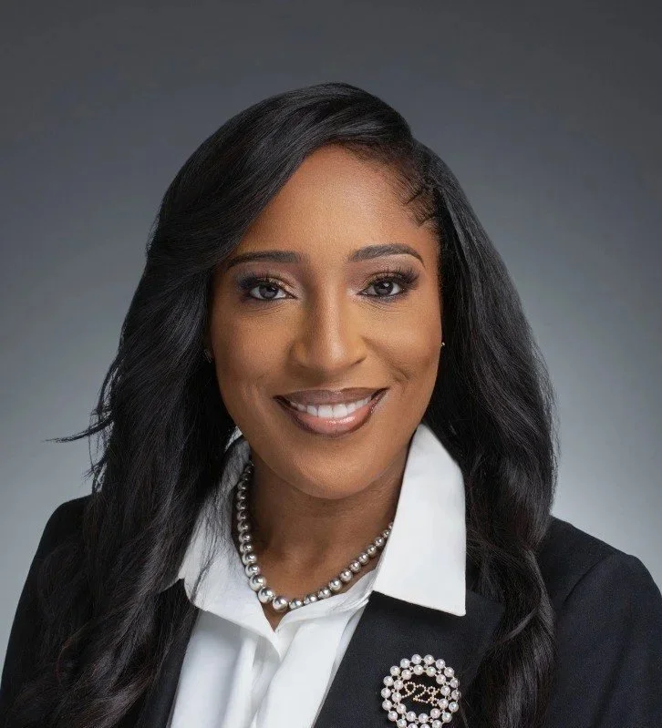Professional portrait of a smiling woman with dark hair, wearing a white collared shirt, a black blazer, a pearl necklace, and a decorative brooch with the number 22.