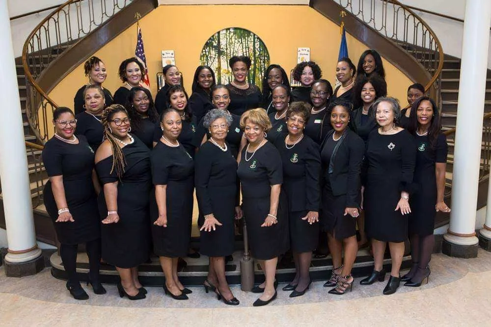 Group of women dressed in black, standing on a staircase inside a building, posing for a photo, with two American flags and a blue flag behind them.