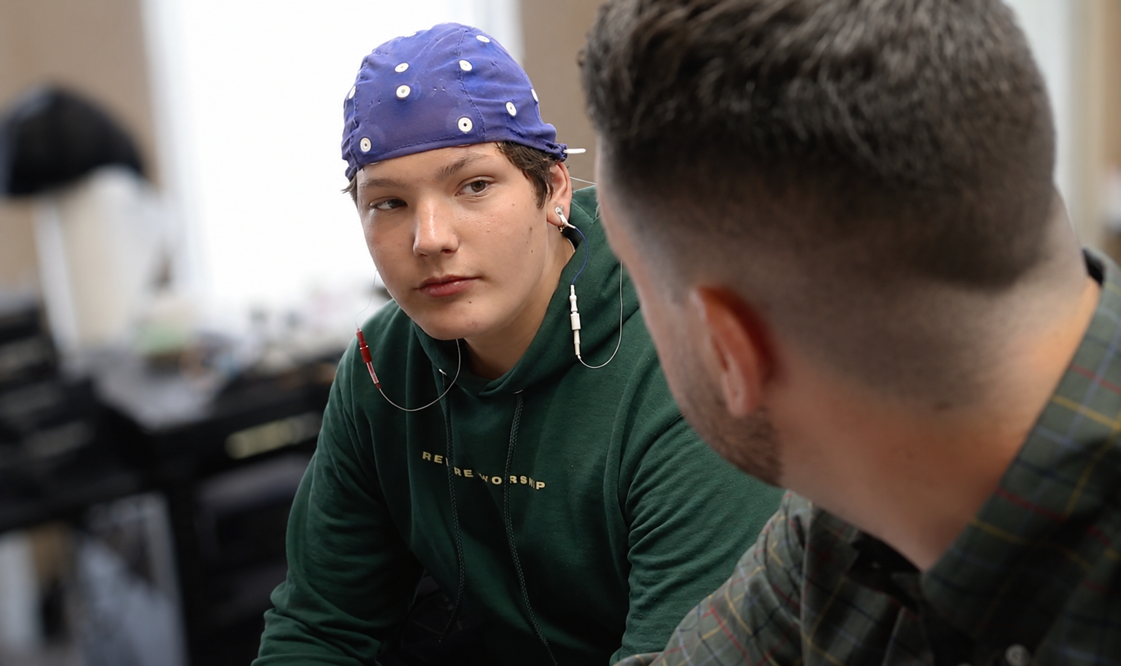 Two young men are engaged in conversation, one wearing a purple bandana with white dots and a green hoodie, the other with short dark hair and a plaid shirt, in a room with blurred objects in the background.