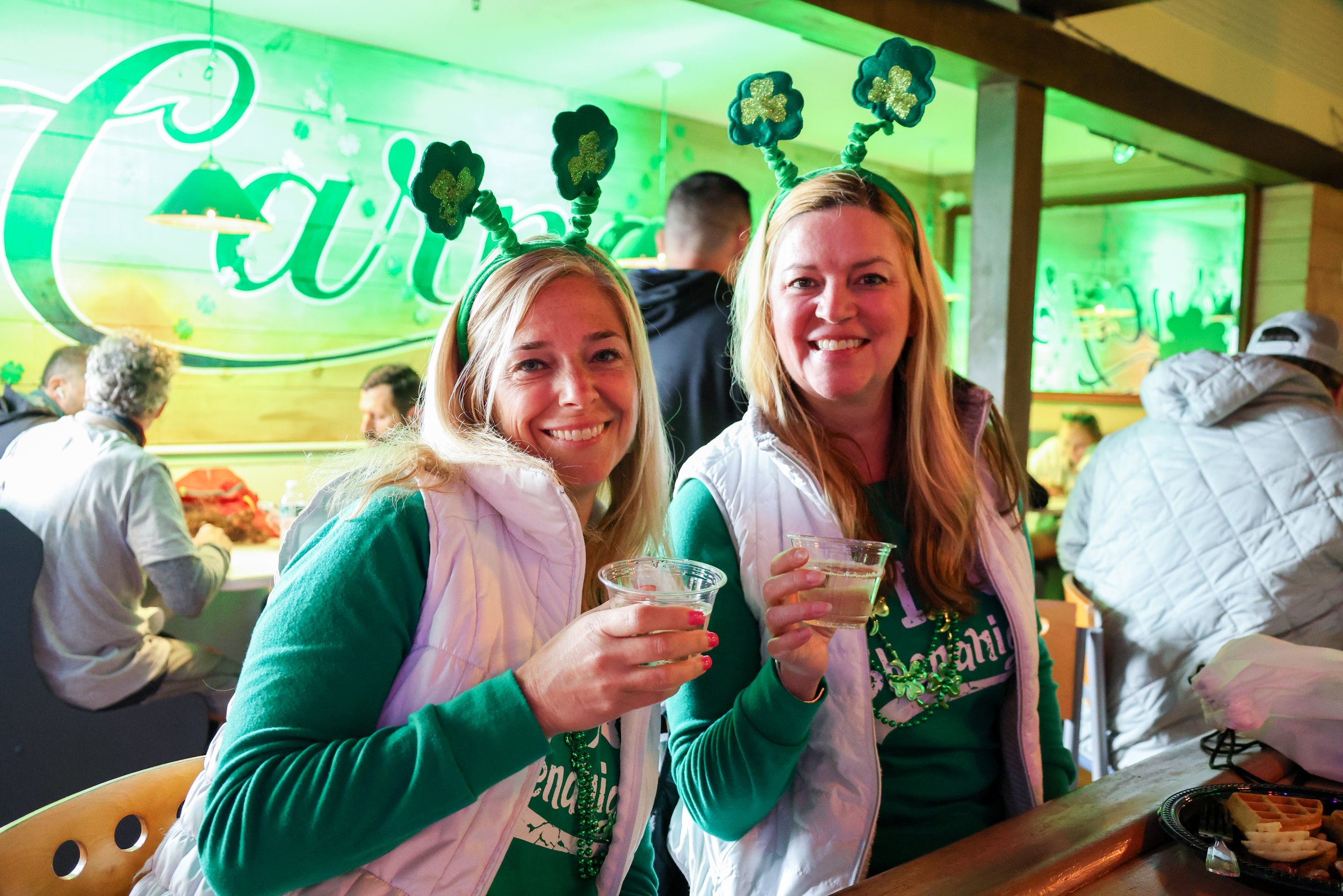 Two women celebrating St. Patrick's Day at a bar, wearing green shirts, green headbands with shamrocks, and holding drinks, with a festive green background.