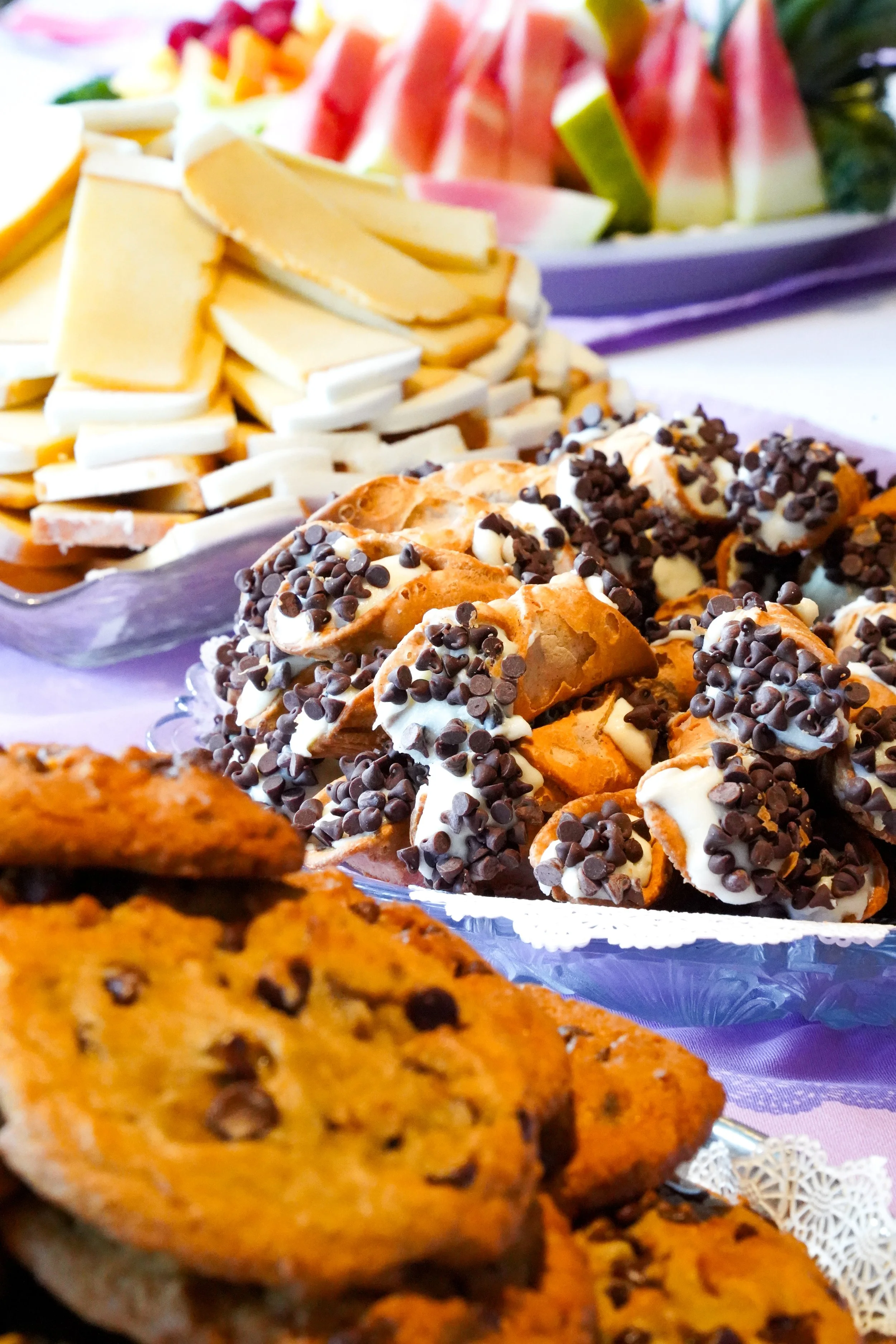 Close-up of assorted desserts including chocolate chip cookies and cannolis topped with chocolate chips, with a platter of fruit in the background.