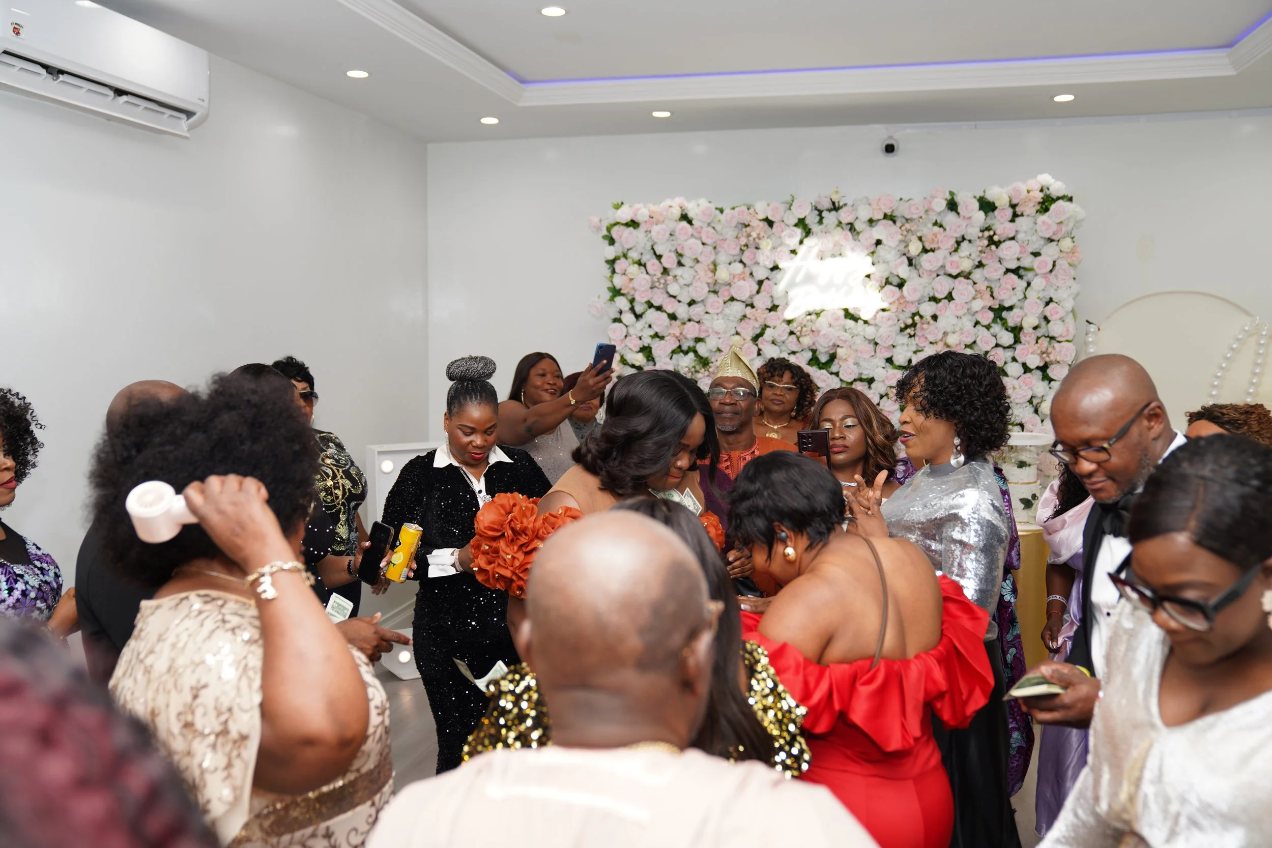 Group of people celebrating at a party with a pink flower wall and a neon sign in background.