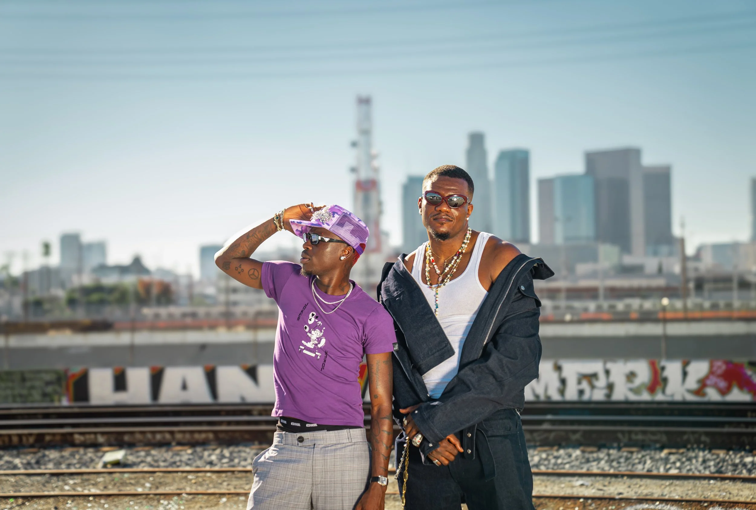 Two stylish men standing on train tracks in front of a city skyline, with graffiti on the wall behind them, both wearing sunglasses and casual streetwear.