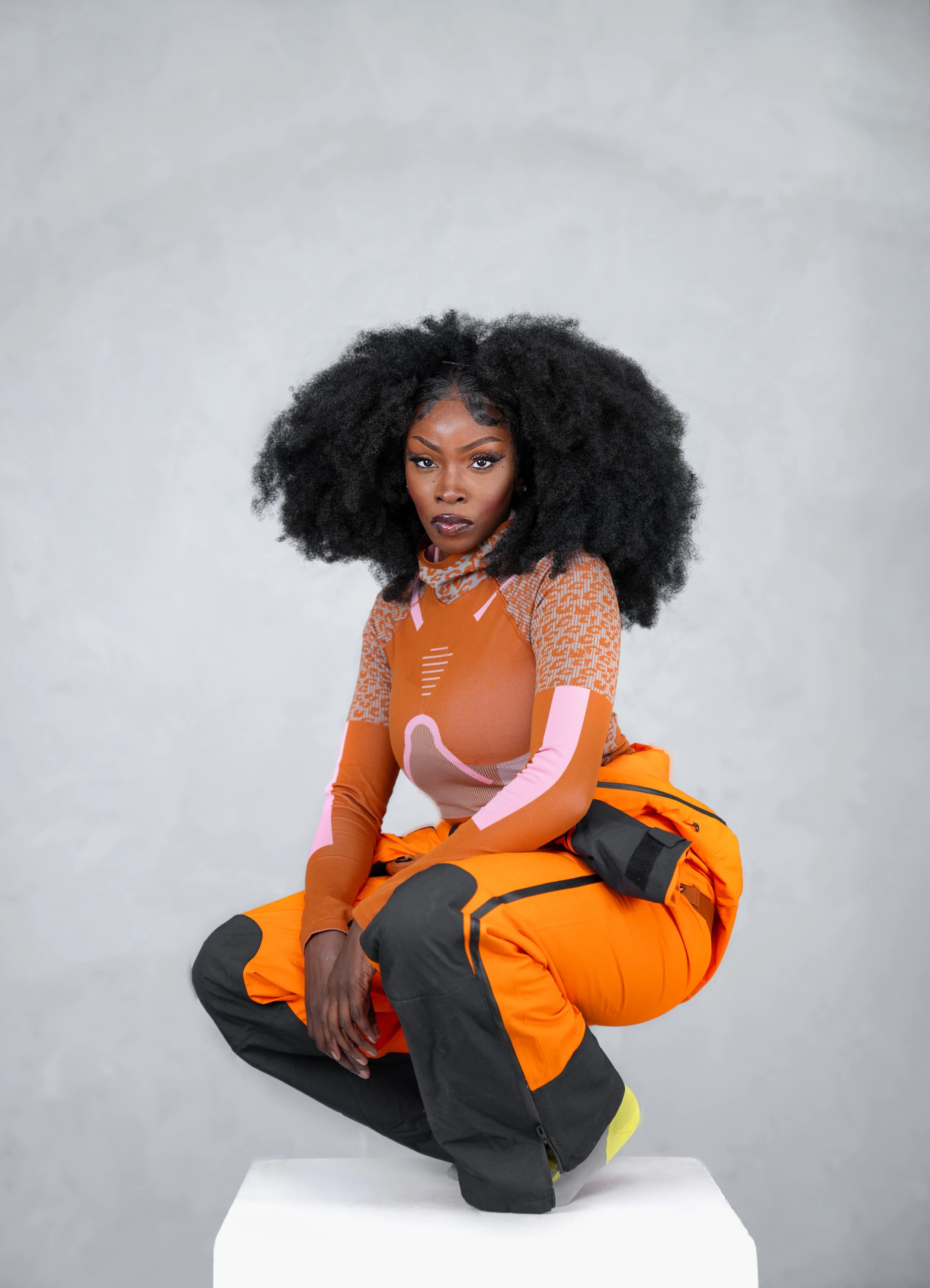A woman with dark skin and large curly black hair crouching on a white platform, wearing a colorful, patterned long-sleeve athletic top and bright orange and black outdoor pants.