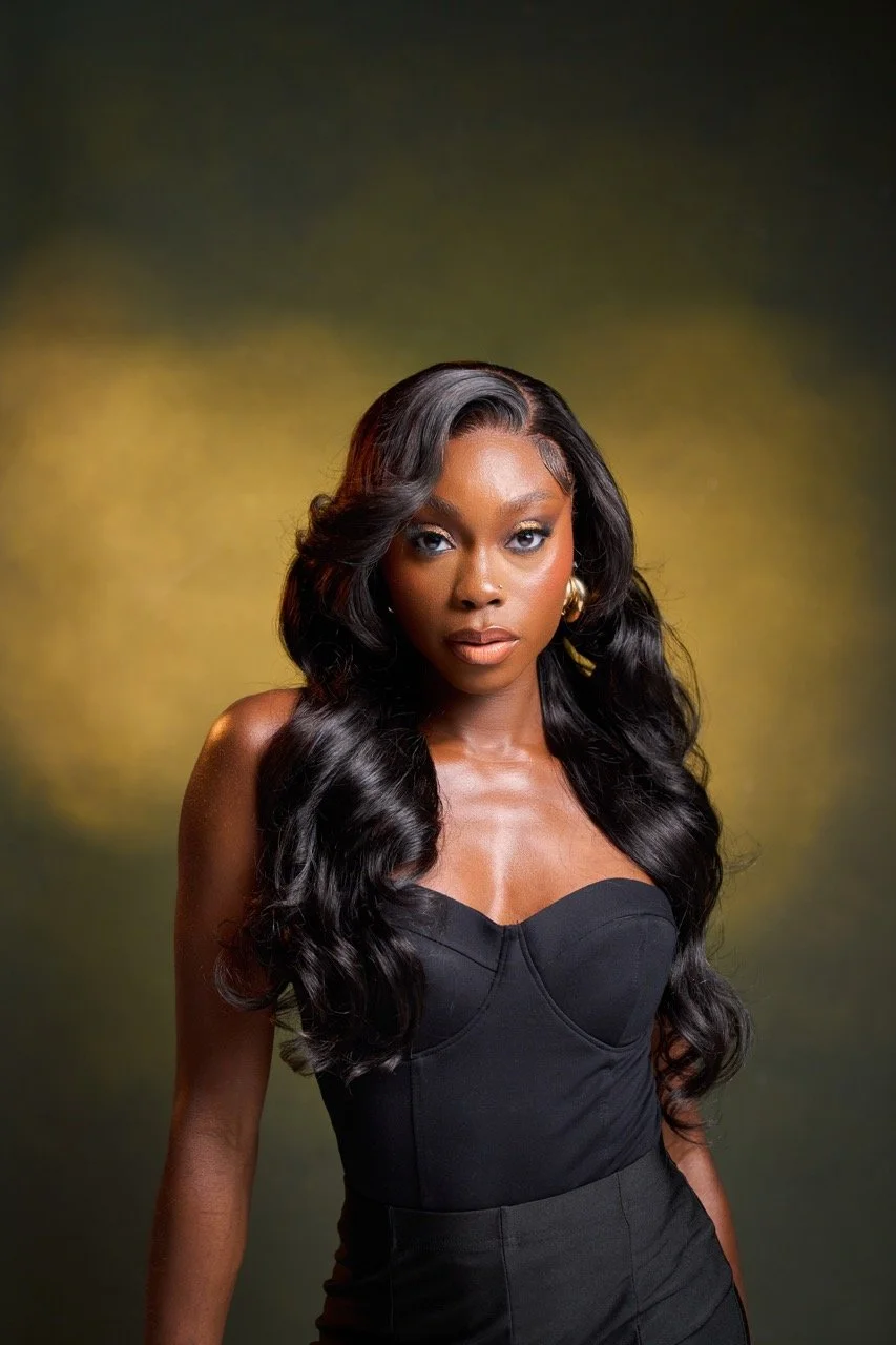 Portrait of a woman with long black wavy hair, wearing a black strapless top and gold earrings, against a dark and yellowish background.