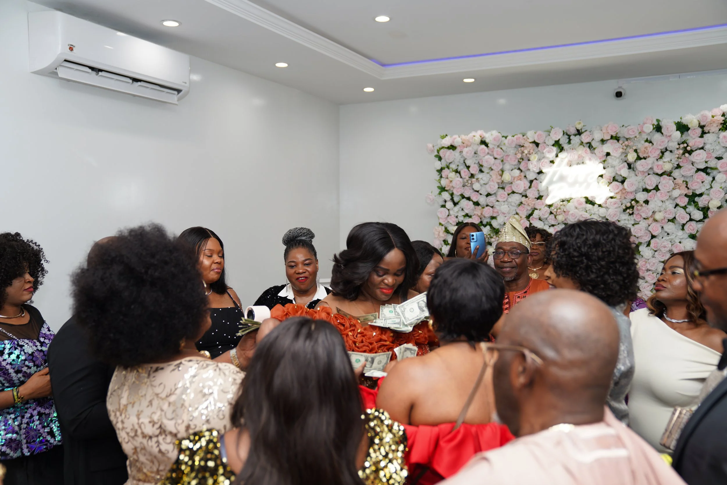 A group of people dressed in formal attire at a celebration, with a woman at the center holding cash, in front of a floral backdrop with a neon sign.