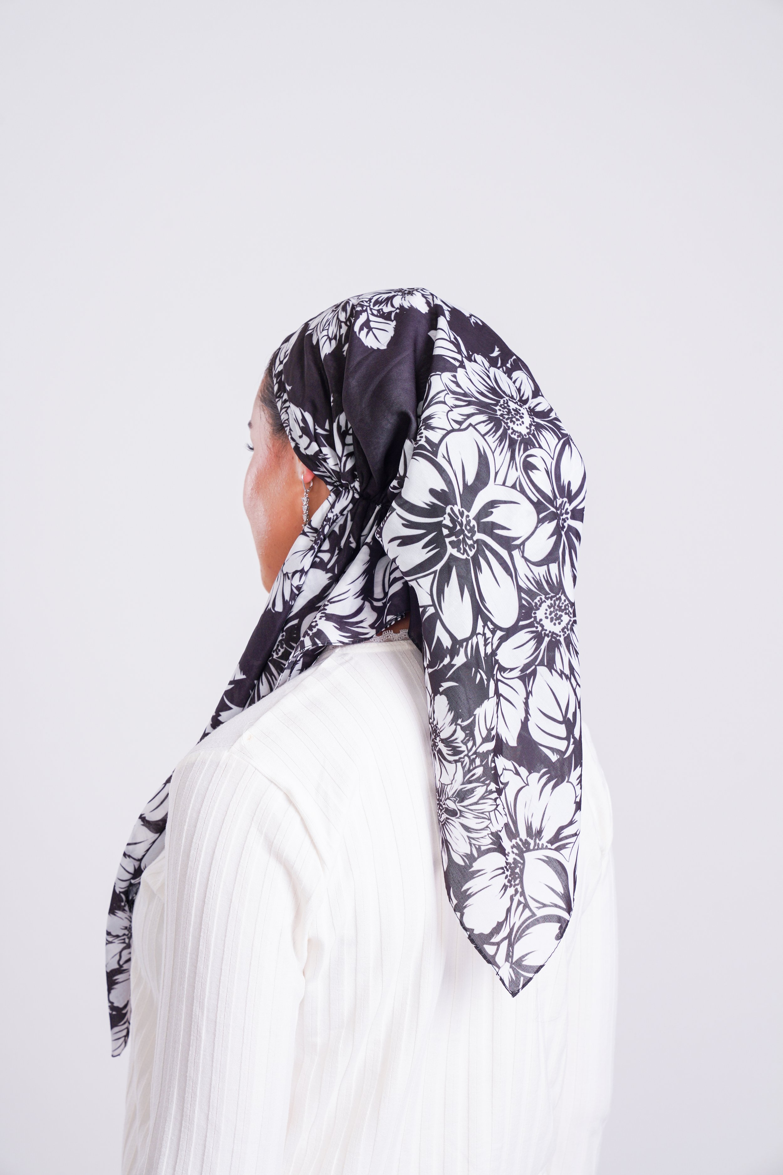 A woman wearing a black and white floral headscarf and a white top, turned to the side against a plain white background.