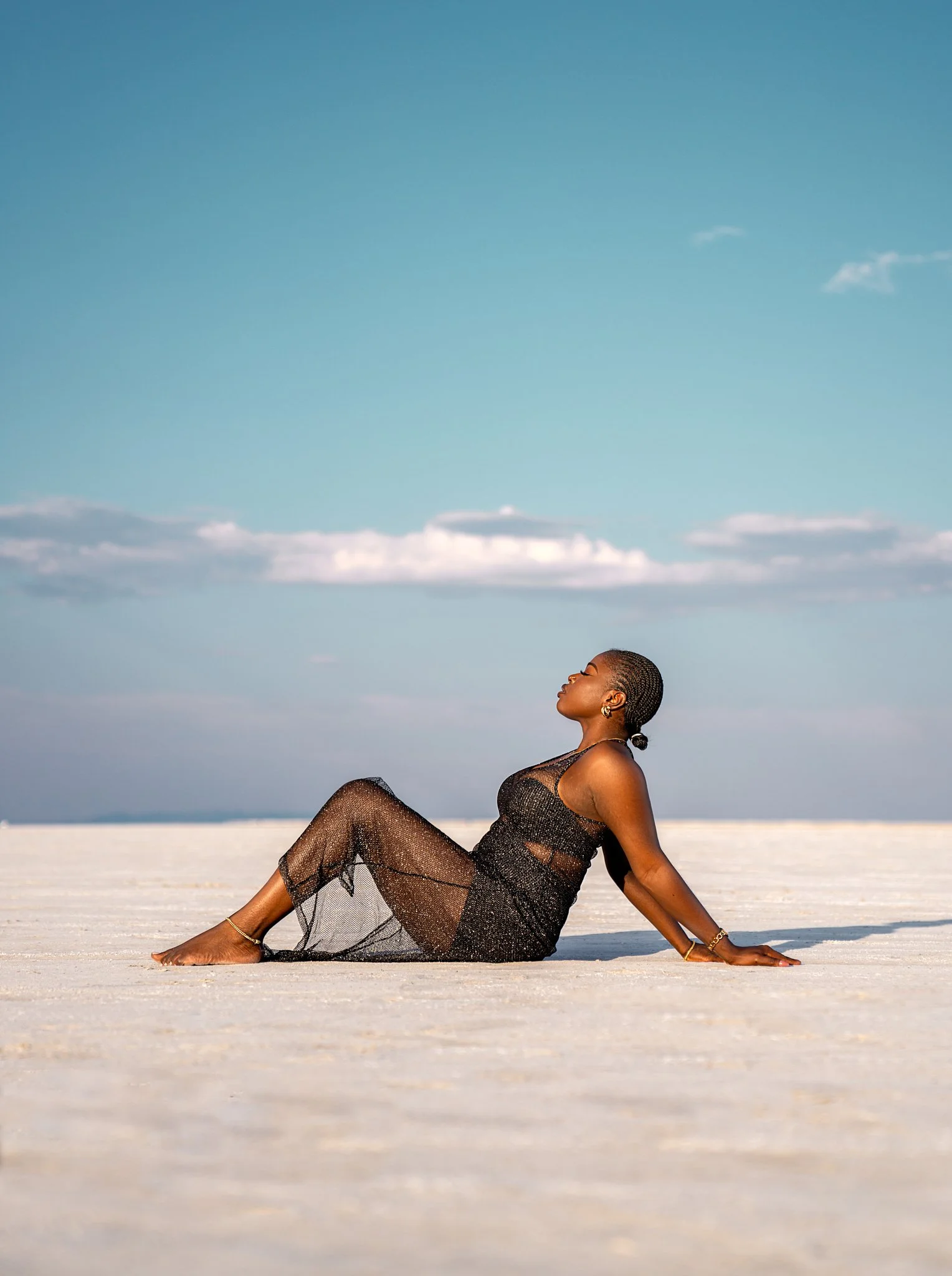 A woman with short hair in a black, semi-sheer dress sitting on a vast white surface under a blue sky with scattered clouds.