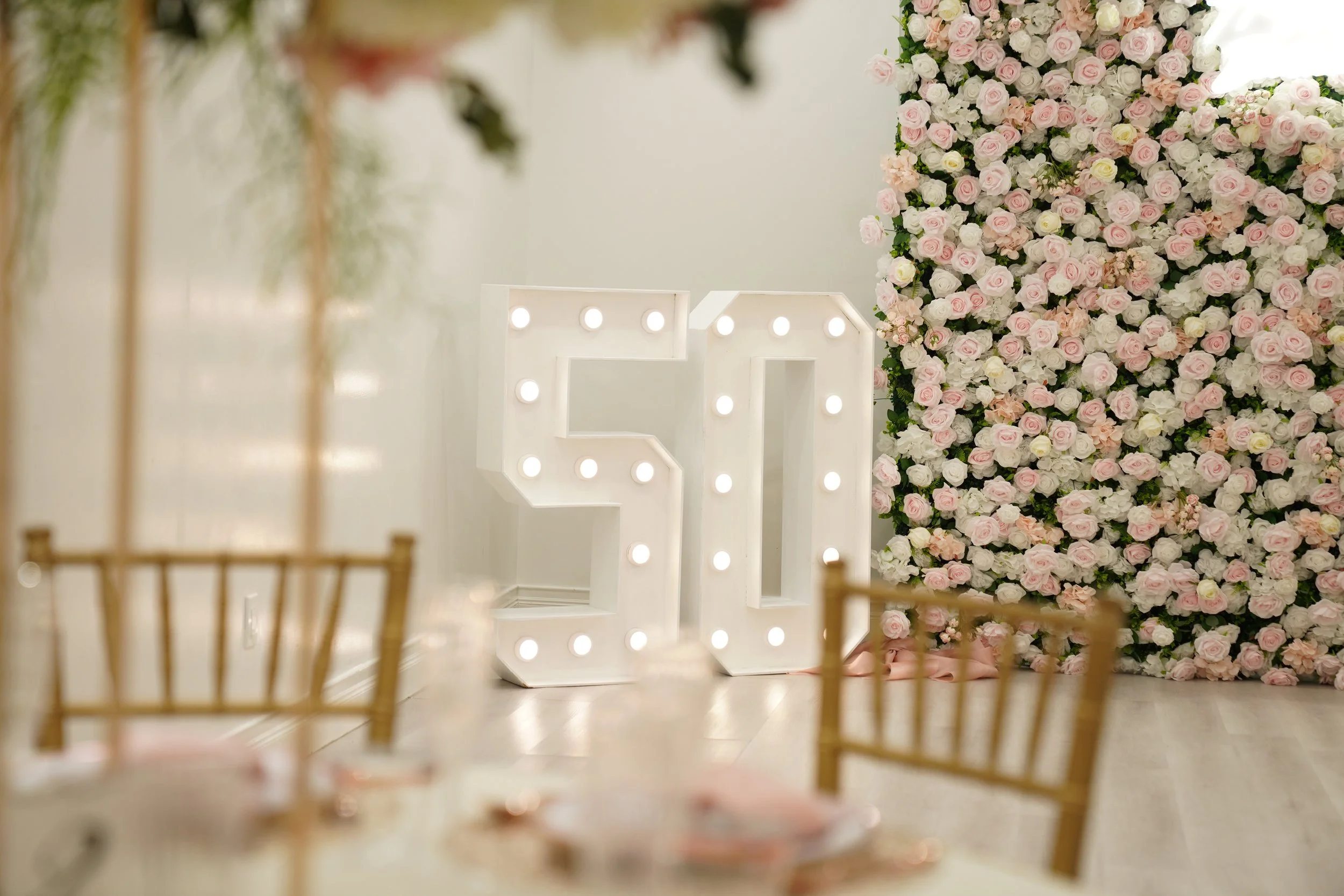 Large illuminated letters 'S' and 'A' in front of a wall of pink and white roses at a celebration or wedding event.