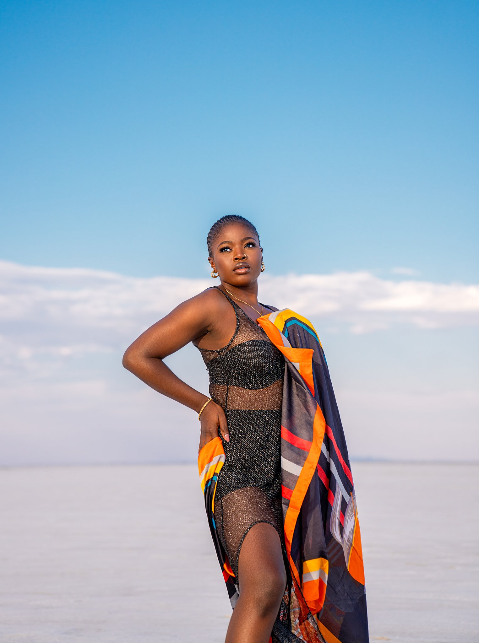 A woman standing on a beach in a black mesh dress with a colorful scarf draped over her shoulder, with a clear blue sky and white clouds in the background.