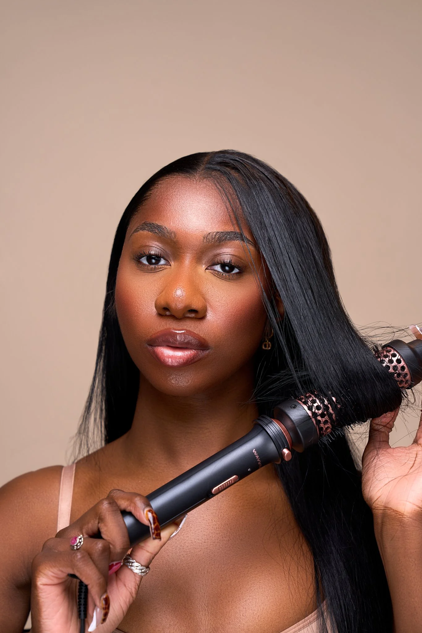 A woman with long dark hair using a curling iron to style her hair in front of a beige background.