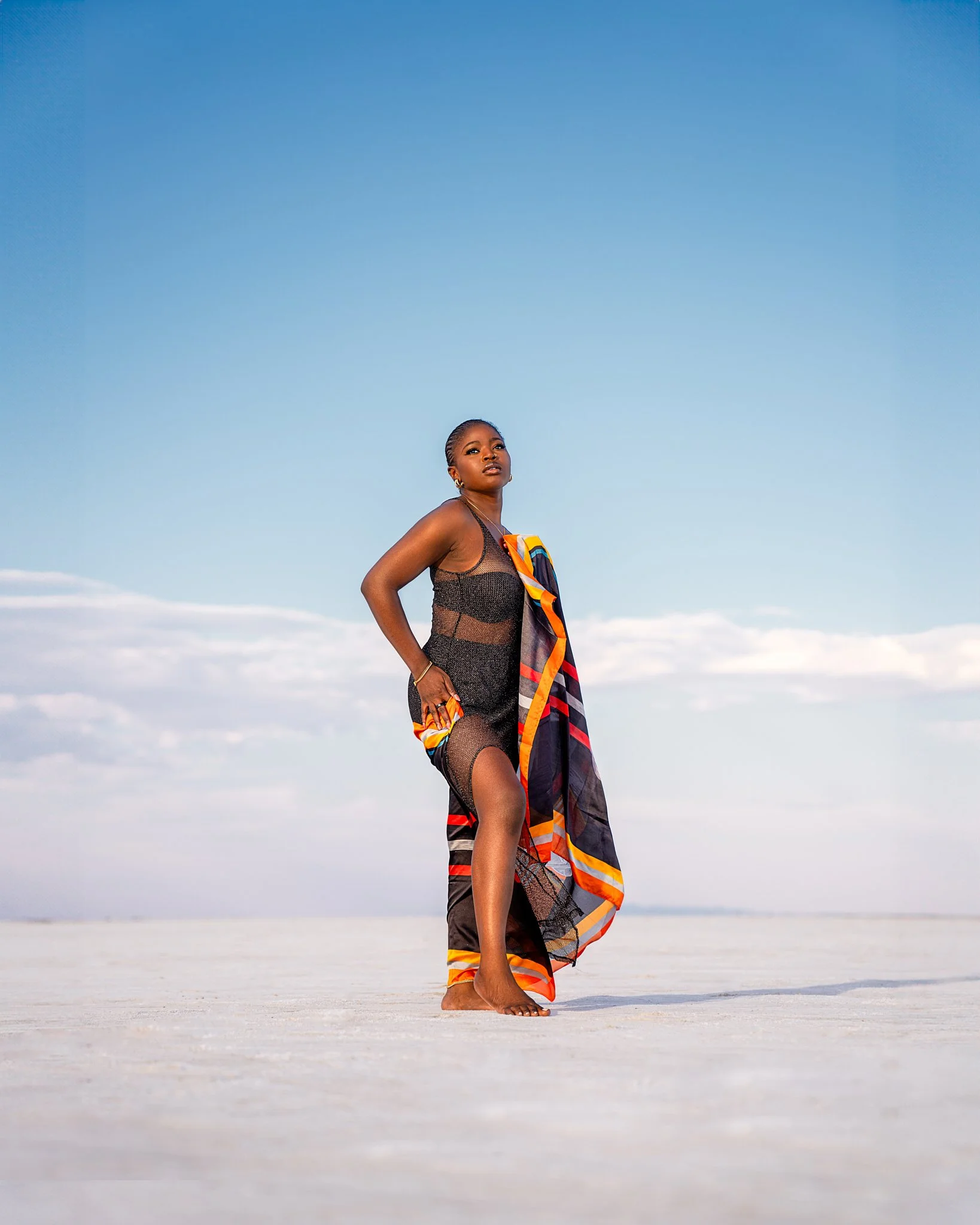 A woman stands barefoot on a flat, white surface with a blue sky and some clouds in the background. She is dressed in black sheer clothing with a colorful scarf draped over her shoulder.