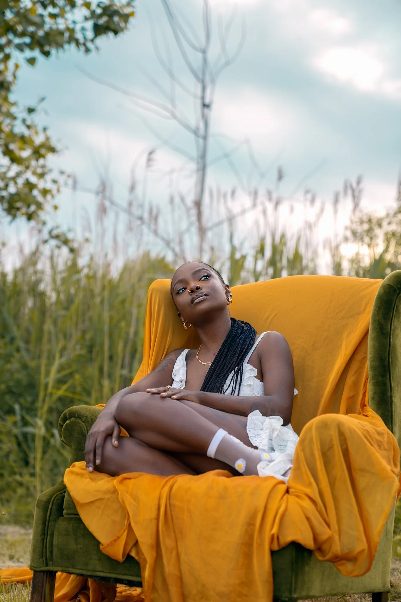 A woman with black braids and gold jewelry sitting on a green armchair with a yellow blanket outdoors in a field of tall grass and trees, with a cloudy sky overhead.