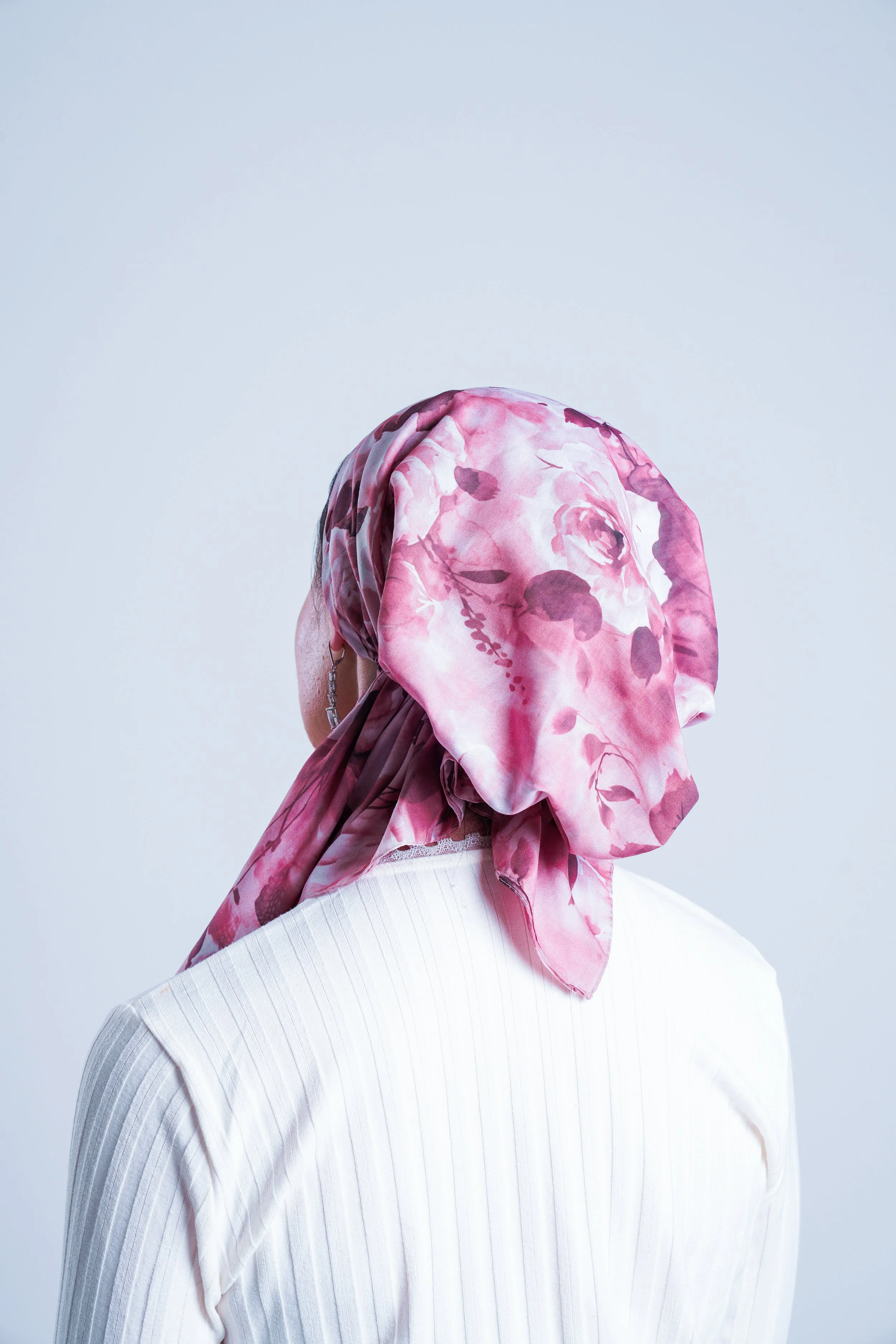 A woman wearing a pink floral headscarf and a white top with vertical stripes, photographed from the side against a plain light background.